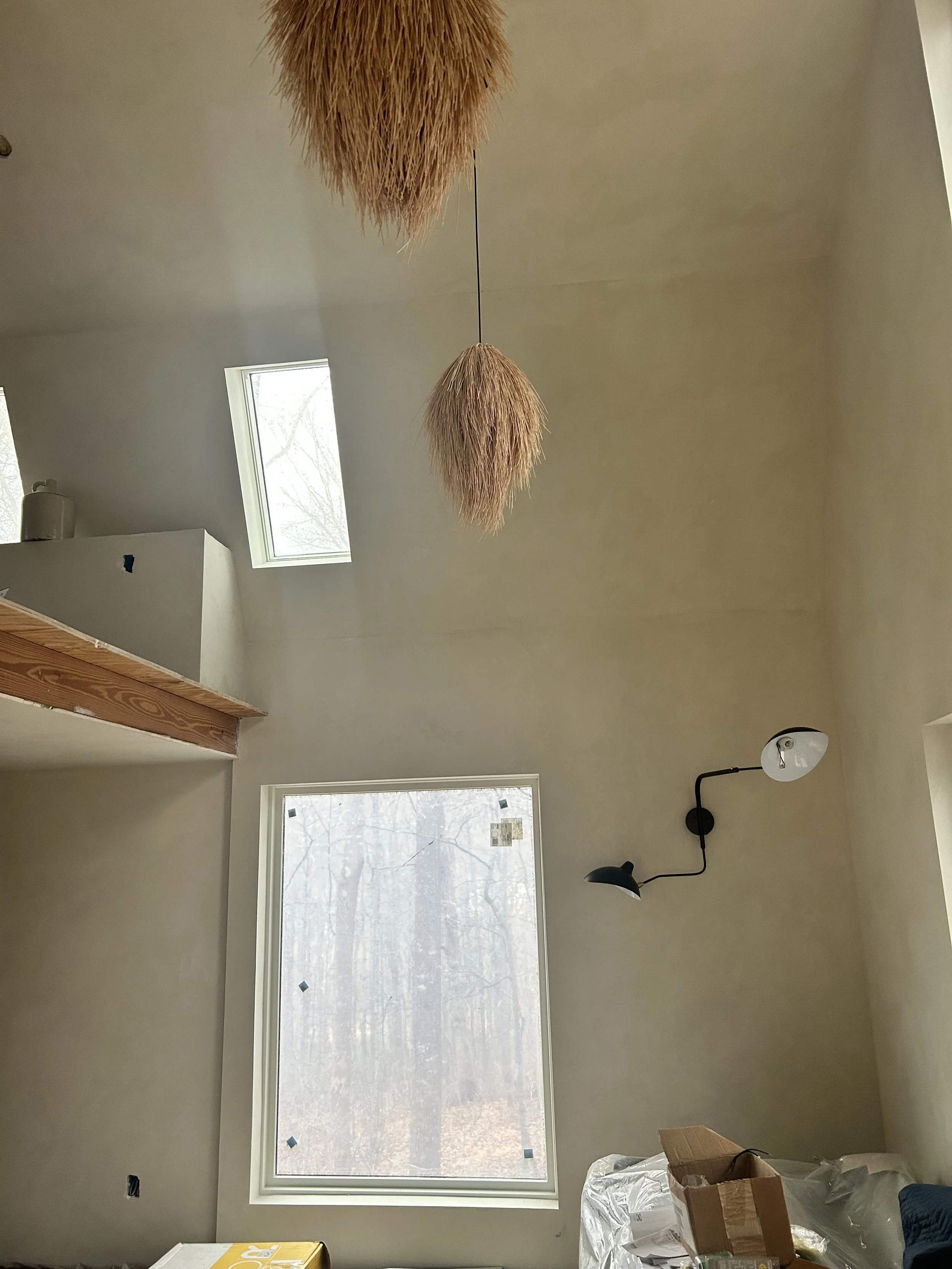 Interior view of a room with a large window, a skylight, and a black wall-mounted lamp. There are some boxes and plastic covering on furniture, suggesting ongoing renovation or moving. The room has high ceilings and minimal decor.
