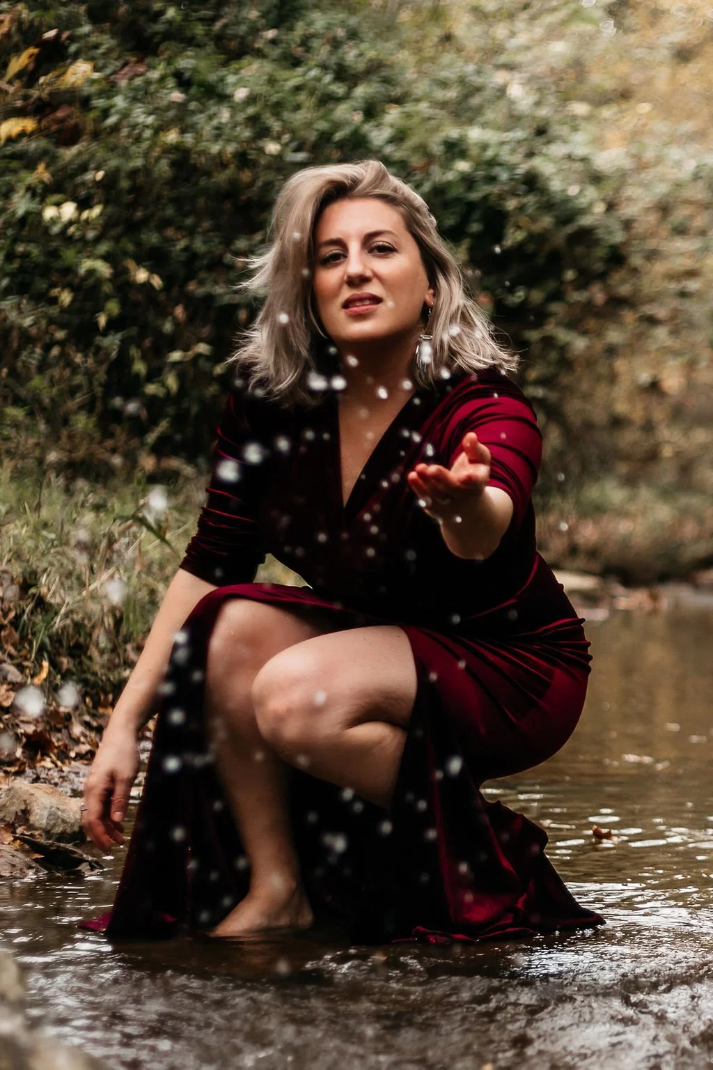 A woman in a red velvet dress crouches in a shallow creek, splashing water towards the camera, surrounded by autumn foliage.