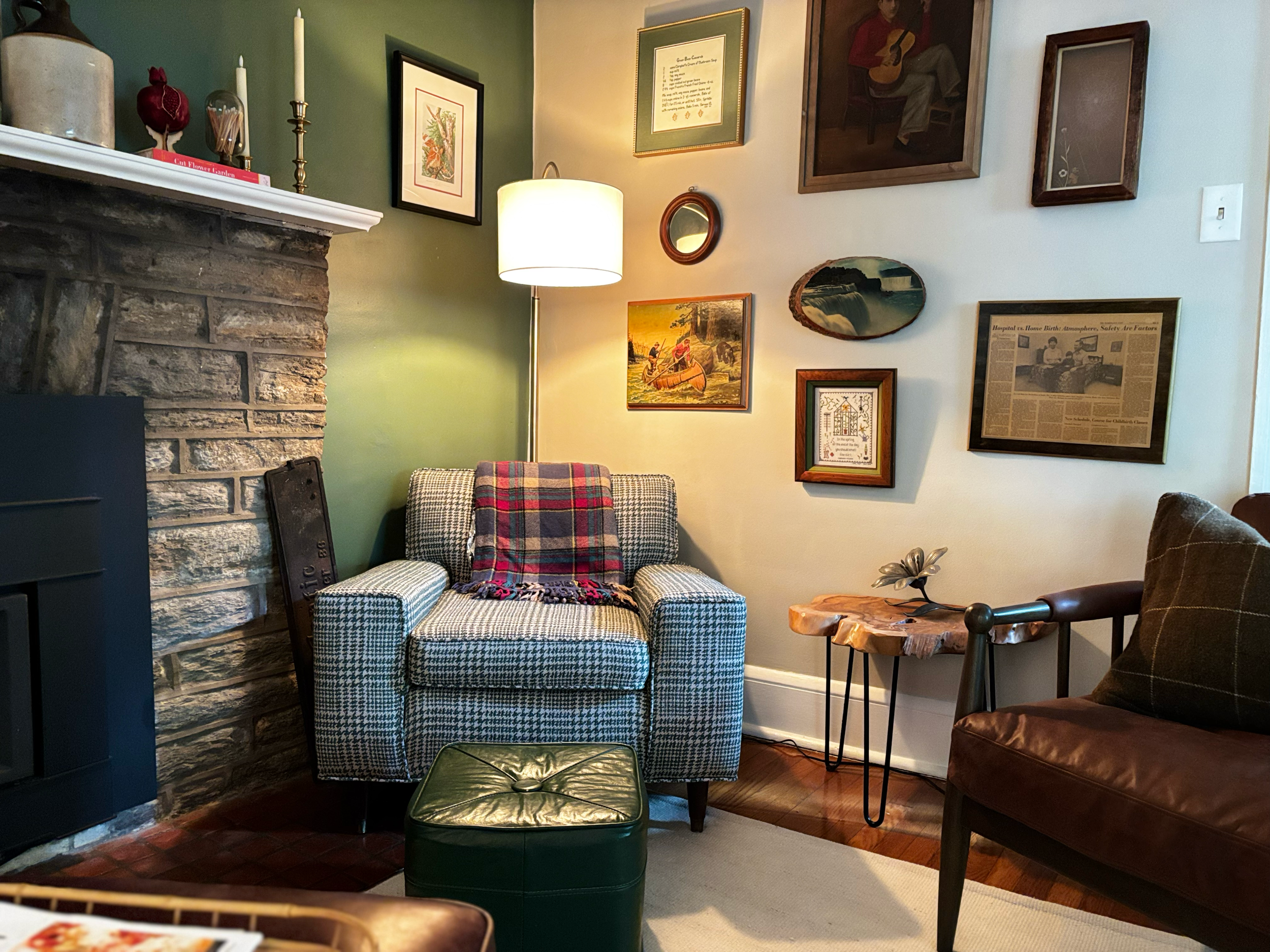 A cozy living room corner with a brick fireplace on the left, a plaid armchair with a blanket, a green ottoman, various framed artworks and photographs on the wall, a lamp providing warm lighting, and a wooden side table with a metallic flower-shaped decor piece.