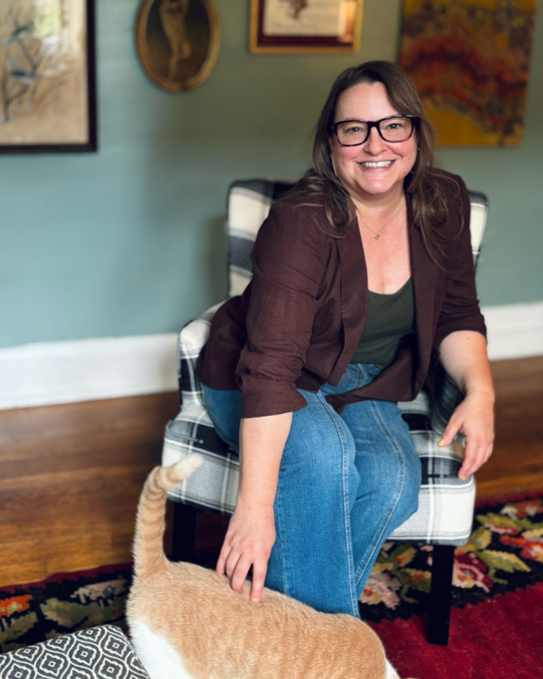A woman with glasses and brown hair sits on a plaid-patterned armchair in a room with framed artwork on the wall. She is smiling and wearing a brown blazer and blue jeans. A ginger cat with a white tip on its tail is in front of her, partially visible.