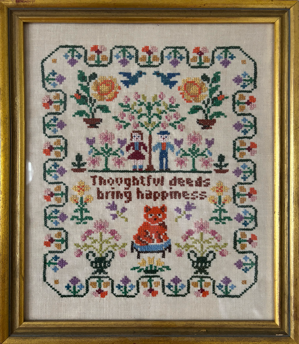 Framed cross-stitch embroidery with colorful flowers, trees, birds, two people, a cat, and the phrase "Thoughtful deeds bring happiness."