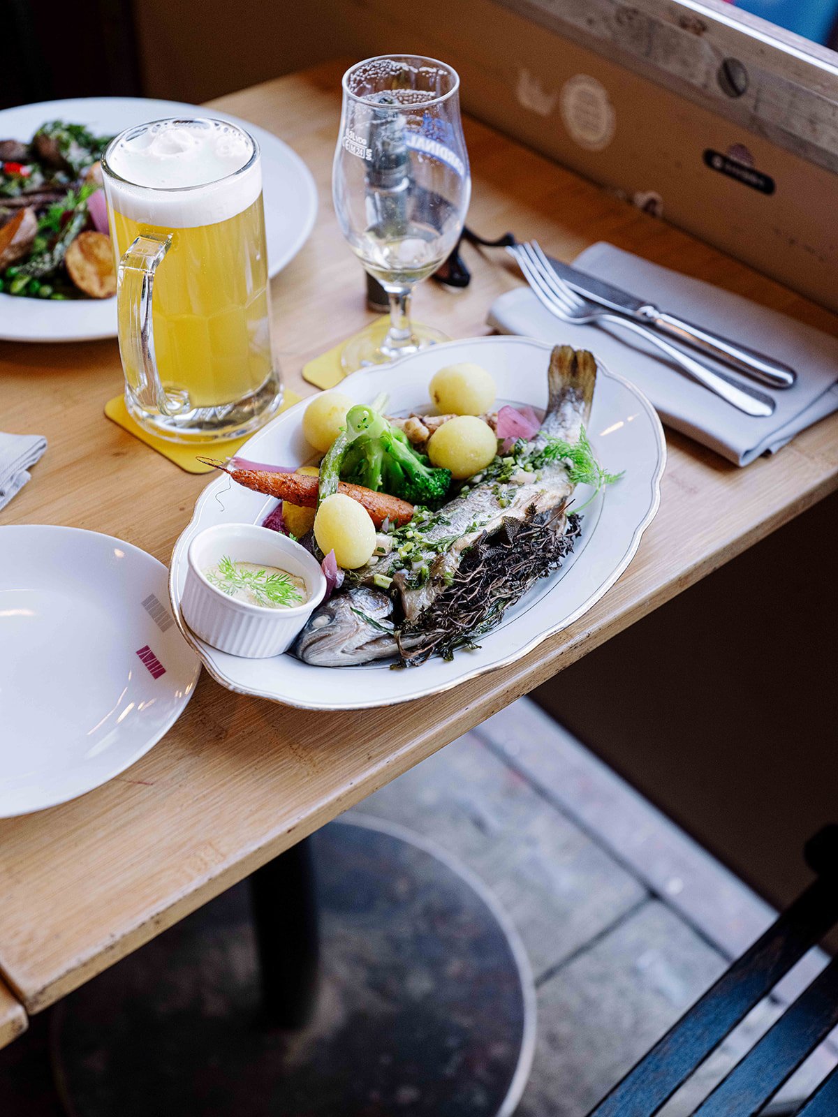 Un plat de poisson servi avec des légumes, des pommes de terre et une sauce, accompagné d'une bière mousseuse, une boisson dans un verre à vin, et une assiette vide à côté. Le repas est présenté sur une table en bois dans un restaurant.