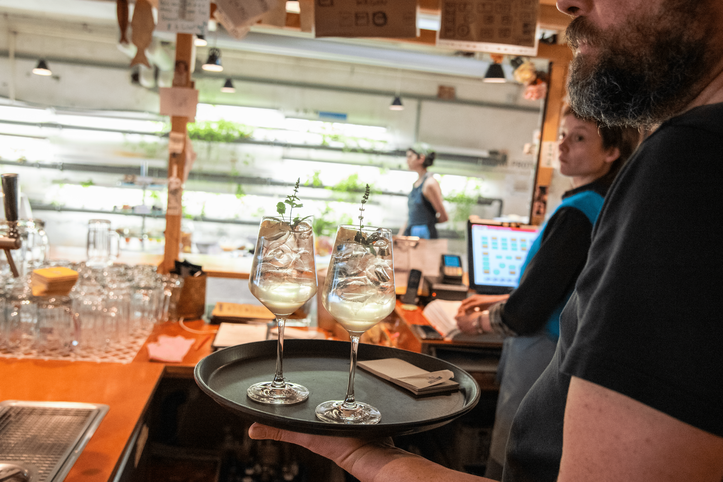 Un serveur présente deux verres à cocktail avec de la glace et de la menthe dans un bar ou un restaurant occupé, avec des clients et du personnel en arrière-plan.