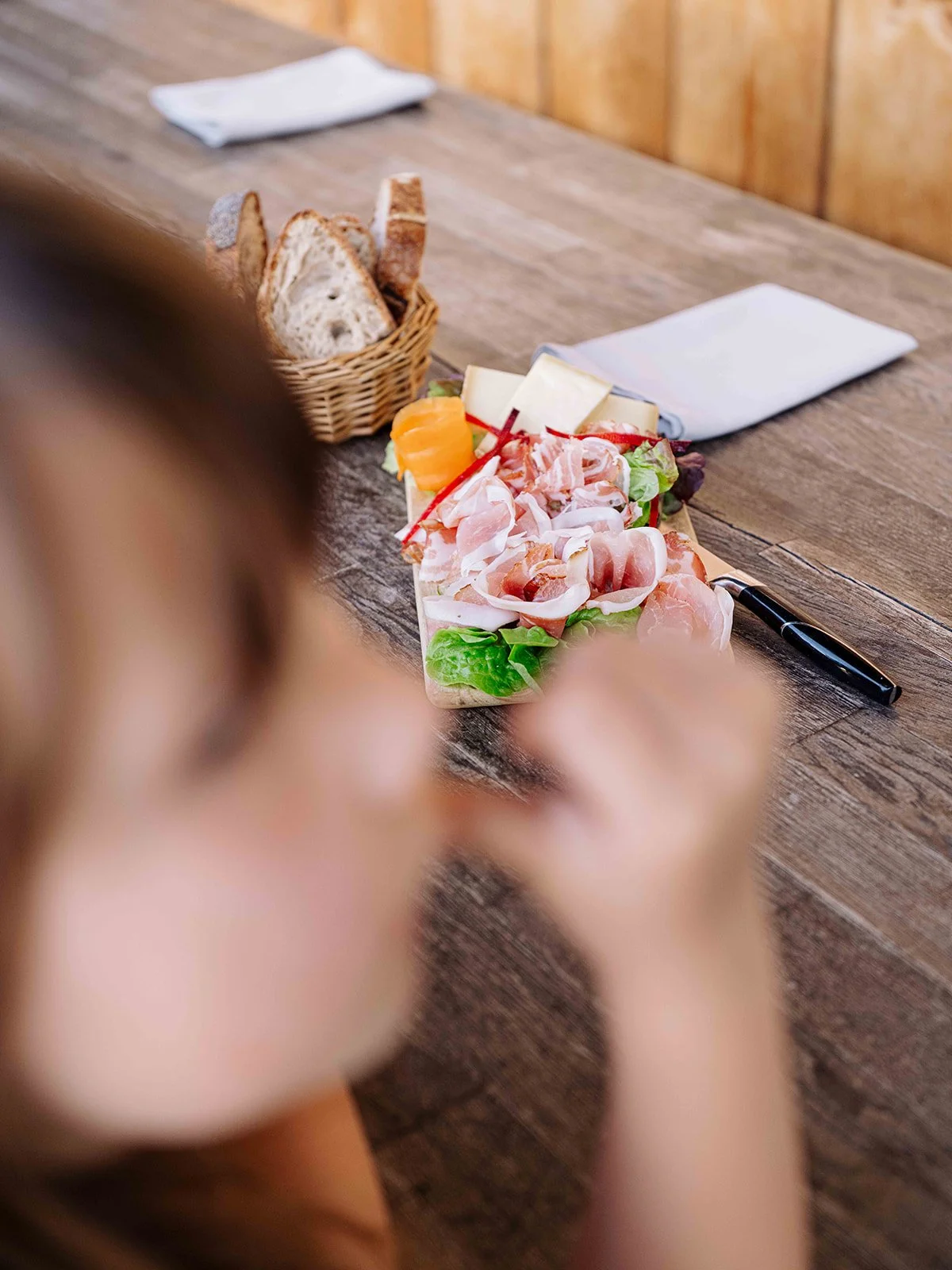 Personne regardant un plateau de charcuterie avec viande, fromage, salade, et fruits, sur une table en bois.