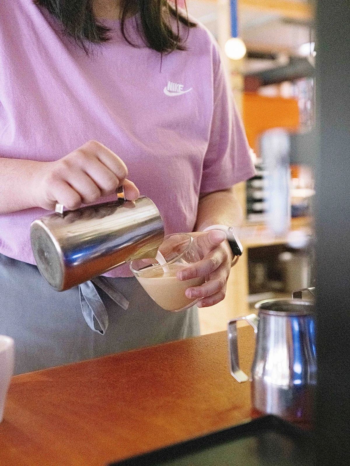 Une personne verse un café ou une boisson chaude dans un verre transparent en cuisine, portant un t-shirt violet Nike.
