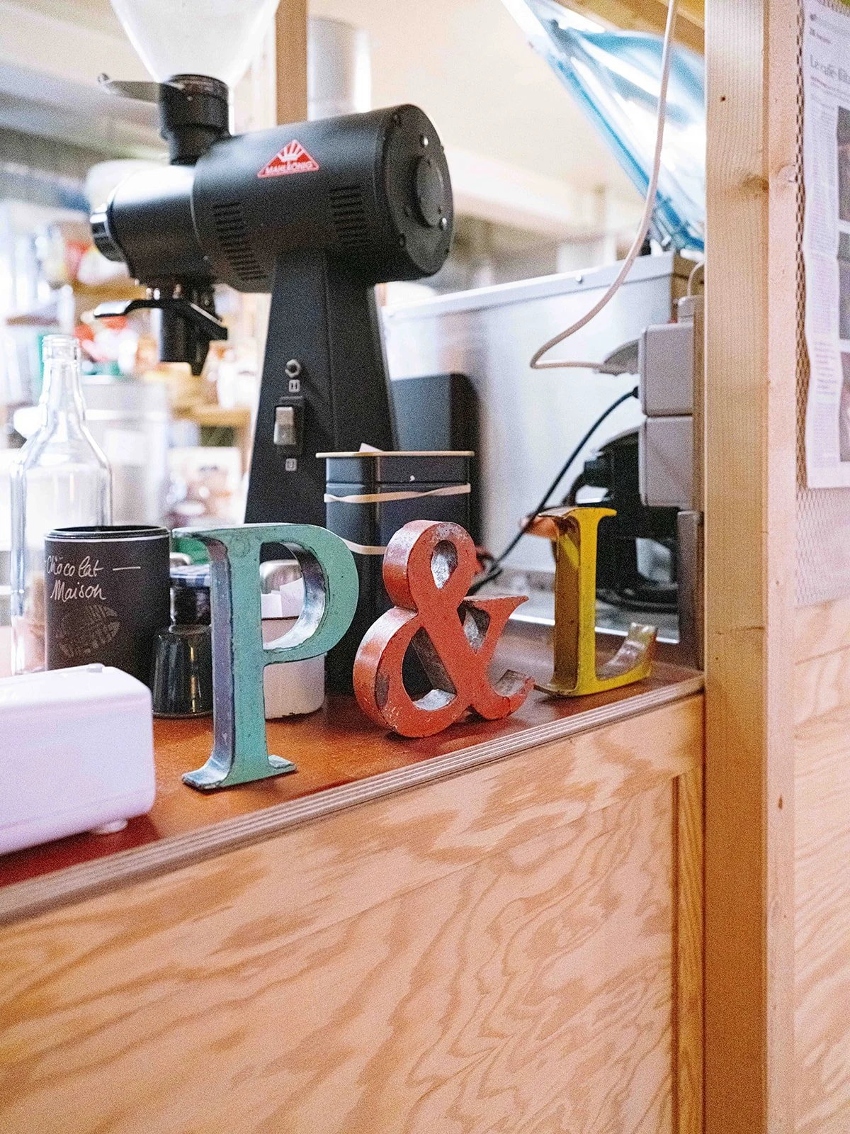 Une étagère en bois avec des lettres décoratives en métal colorées P, &, L, une cafetière noir, une tasse en noir, une bouteille en verre, et un cafetière électrique.