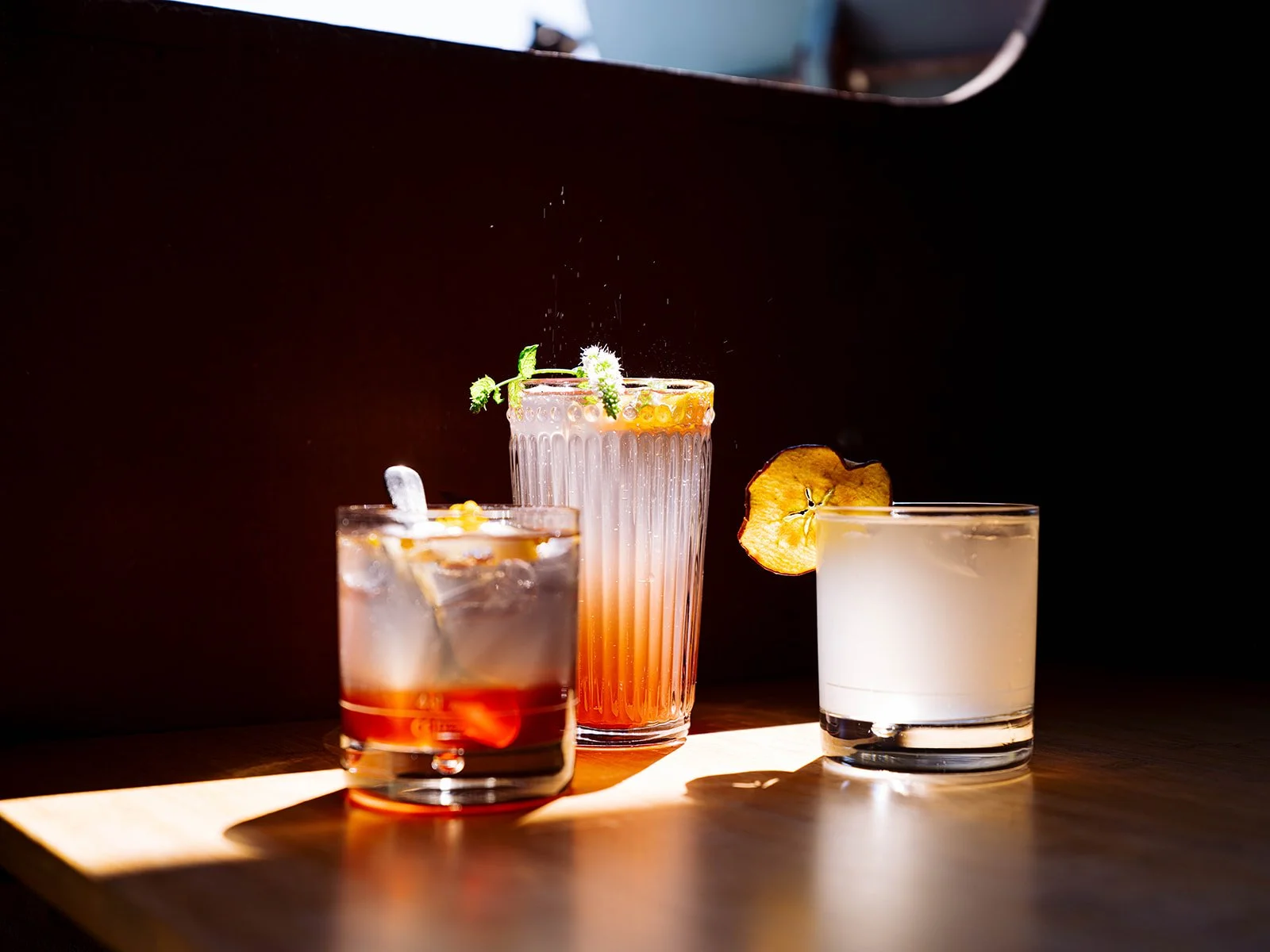 Trois cocktails différents sur une table en bois, avec des décorations de fruits et des fleurs, éclairés par une lumière naturelle.