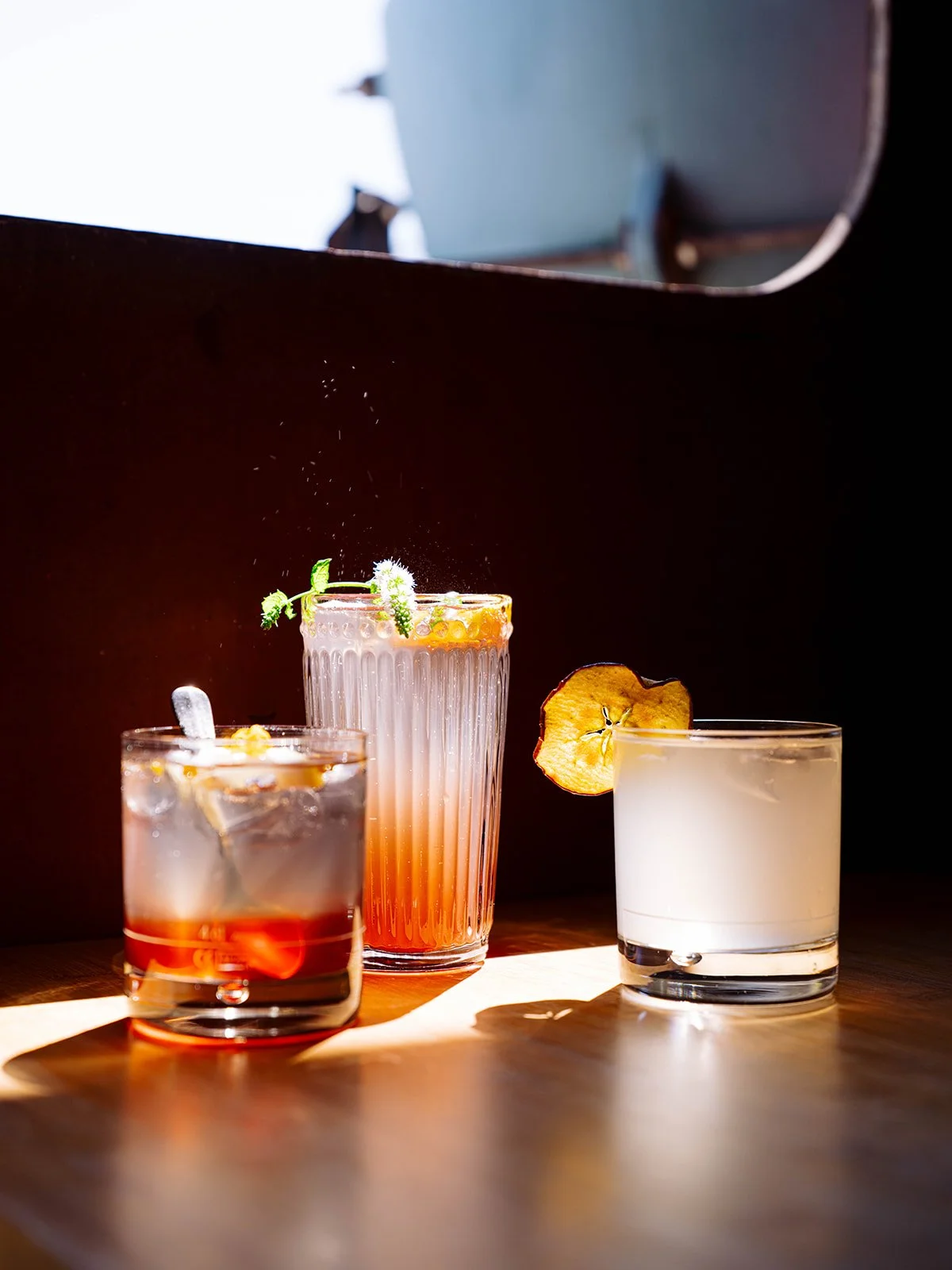 Trois cocktails colorés sur une table en bois, avec un fond sombre. Le premier à gauche est un verre court avec un cocktail rouge et de la glace. Le centre est un grand verre avec un cocktail rose pâle, décoré de feuilles de menthe et d'une tranches 