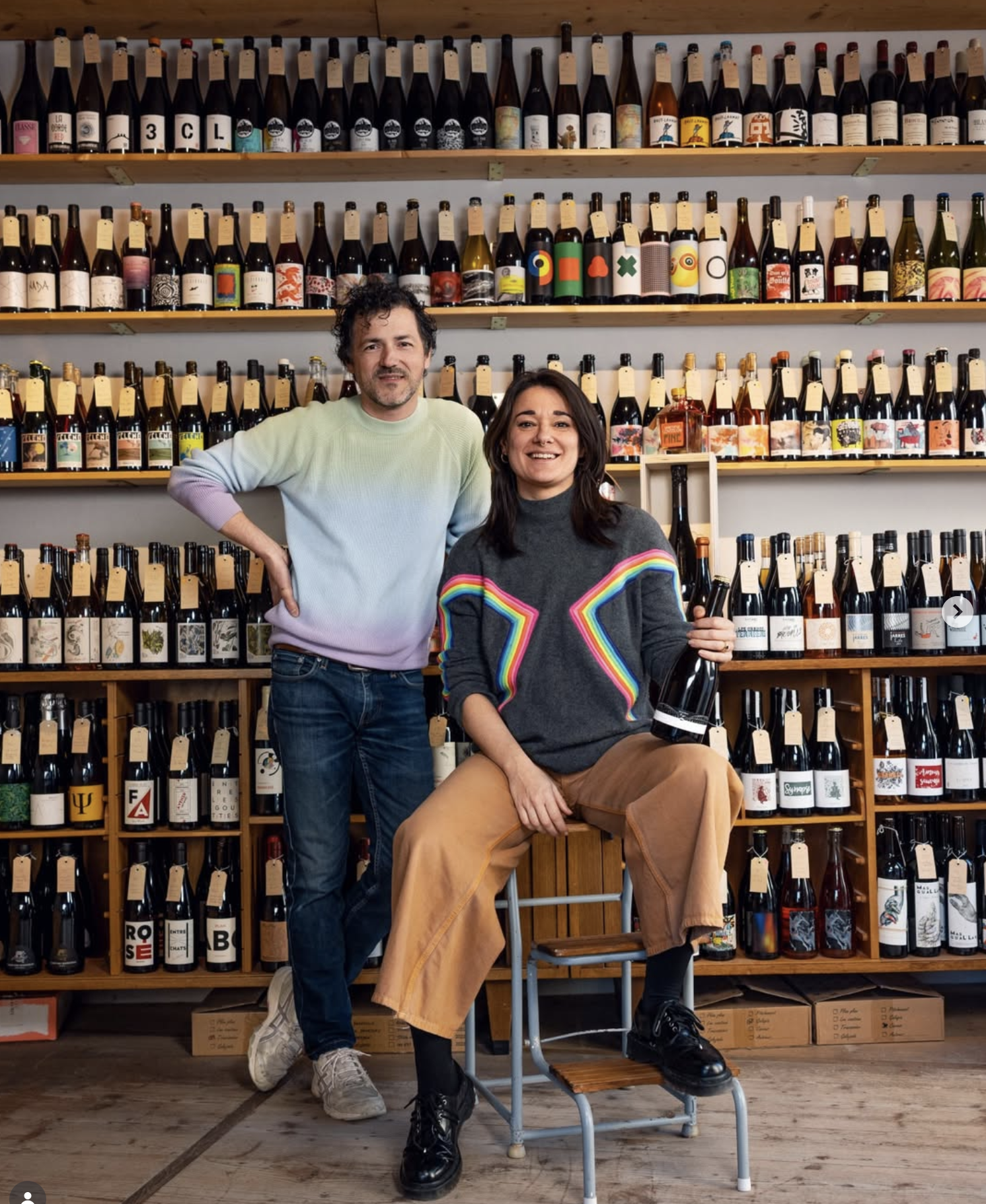 Deux personnes dans une cave à vin. L'une se tient debout avec une main sur la hanche, l'autre est assise sur une chaise, tenant une bouteille de vin. En arrière-plan, étagères remplies de bouteilles de vin aux étiquettes colorées.