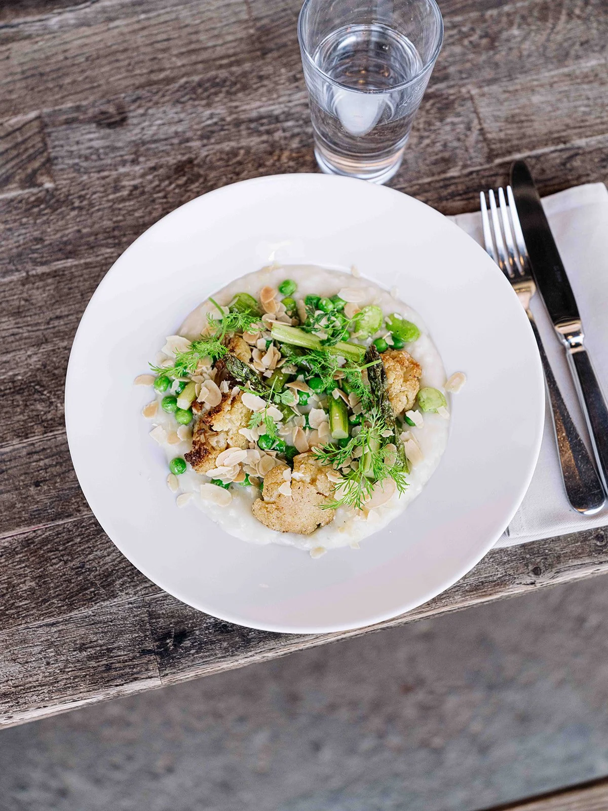 Assiette de florets de chou-fleur, asperges, petits pois, herbes fraîches, amandes effilées sur un lit de sauce blanche, accompagnée d'un verre d'eau dans un restaurant