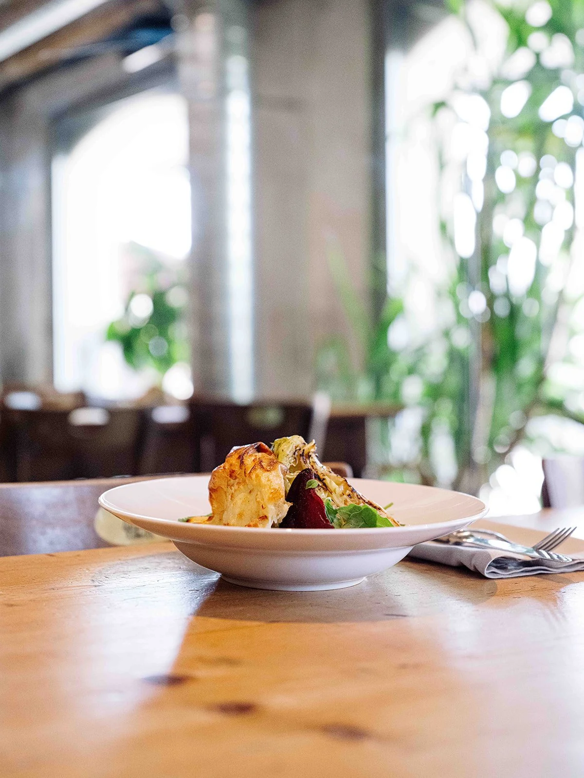 Un plat de nourriture chaud avec des légumes et une portion de gratin, placé sur une table en bois dans un restaurant avec un intérieur lumineux et verdoyant.