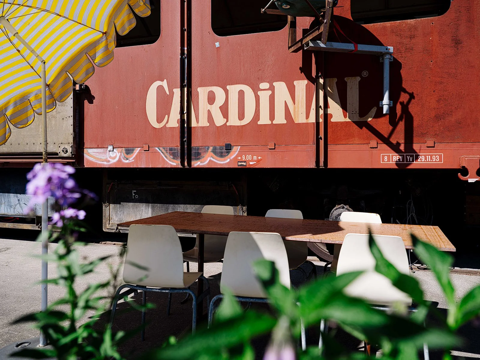 Une table en bois entourée de chaises blanches dans un espace extérieur avec un train rouge marqué 'Cardinal' en arrière-plan, décoré avec un parasol jaune rayé et des fleurs en premier plan.
