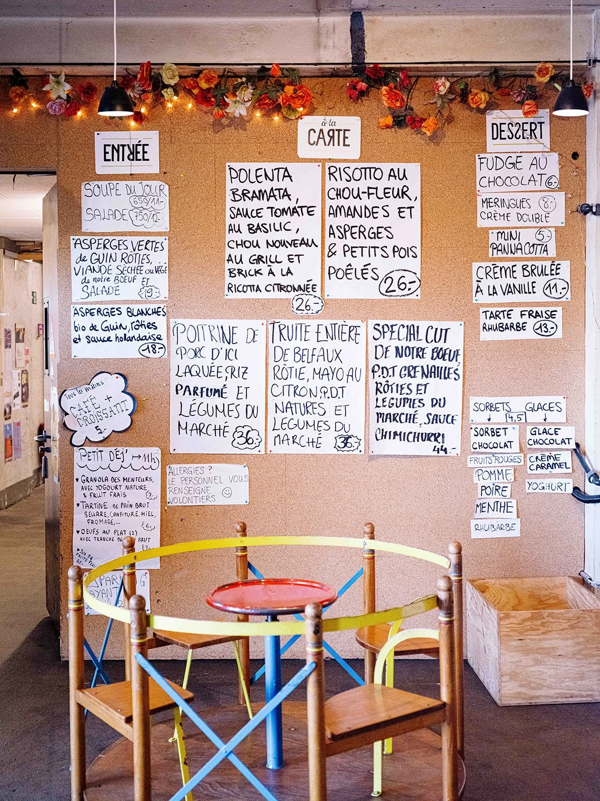 Panneaux de menu écrits à la main affichant des plats et desserts en français, décorés de guirlandes de fleurs artificielles, dans un restaurant. Chambres avec petite table centrale et chaises colorées en bois.