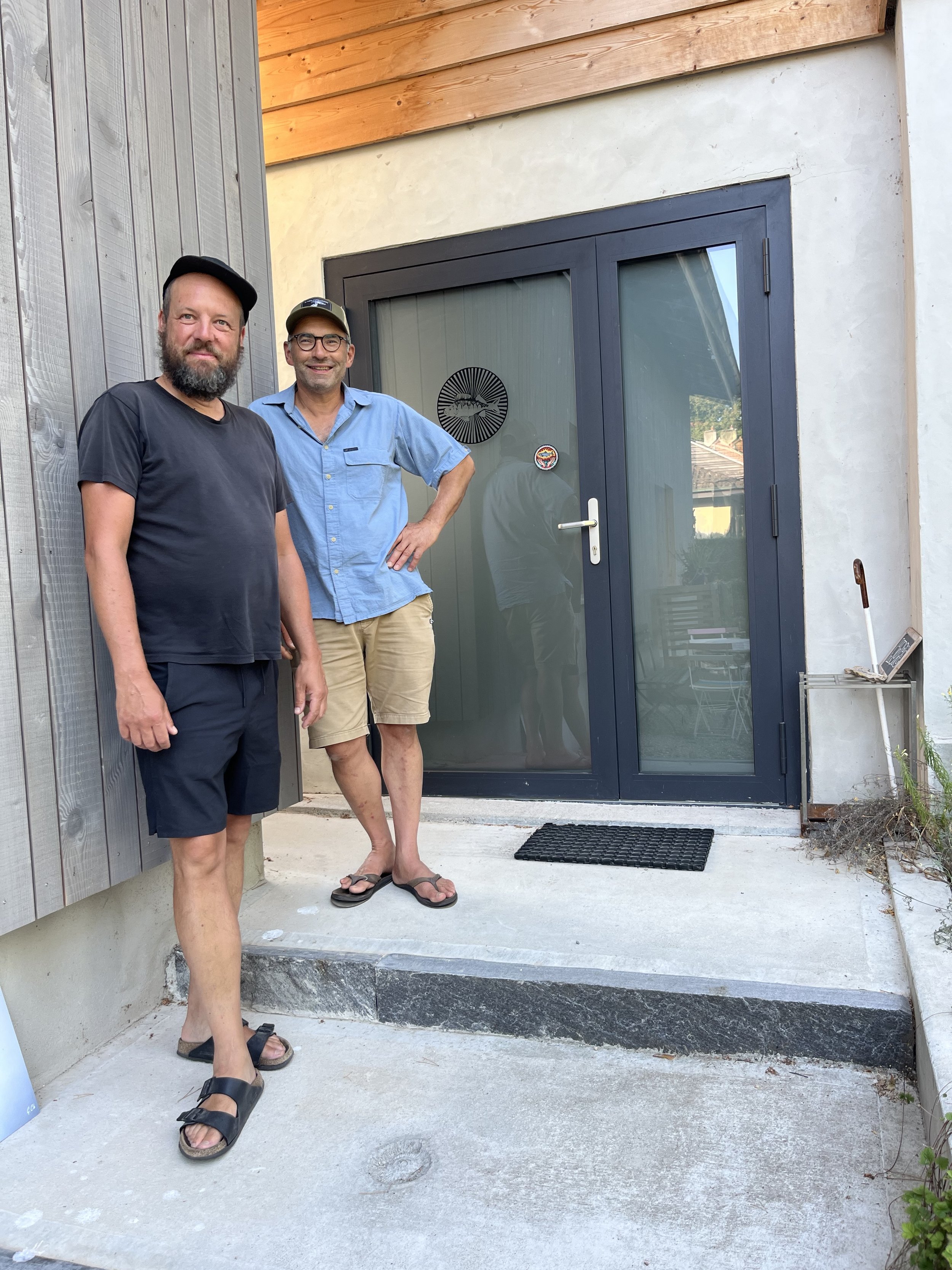 Deux hommes debout devant une porte vitrée, un porte-clés suspendu à la porte et un petit panneau à côté. La porte est encadrée d'une façade en béton et en bois. Les hommes portent des vêtements décontractés, l'un en t-shirt noir et l'autre en chemise bleue, et portent des sandales ou des chaussures ouvertes.