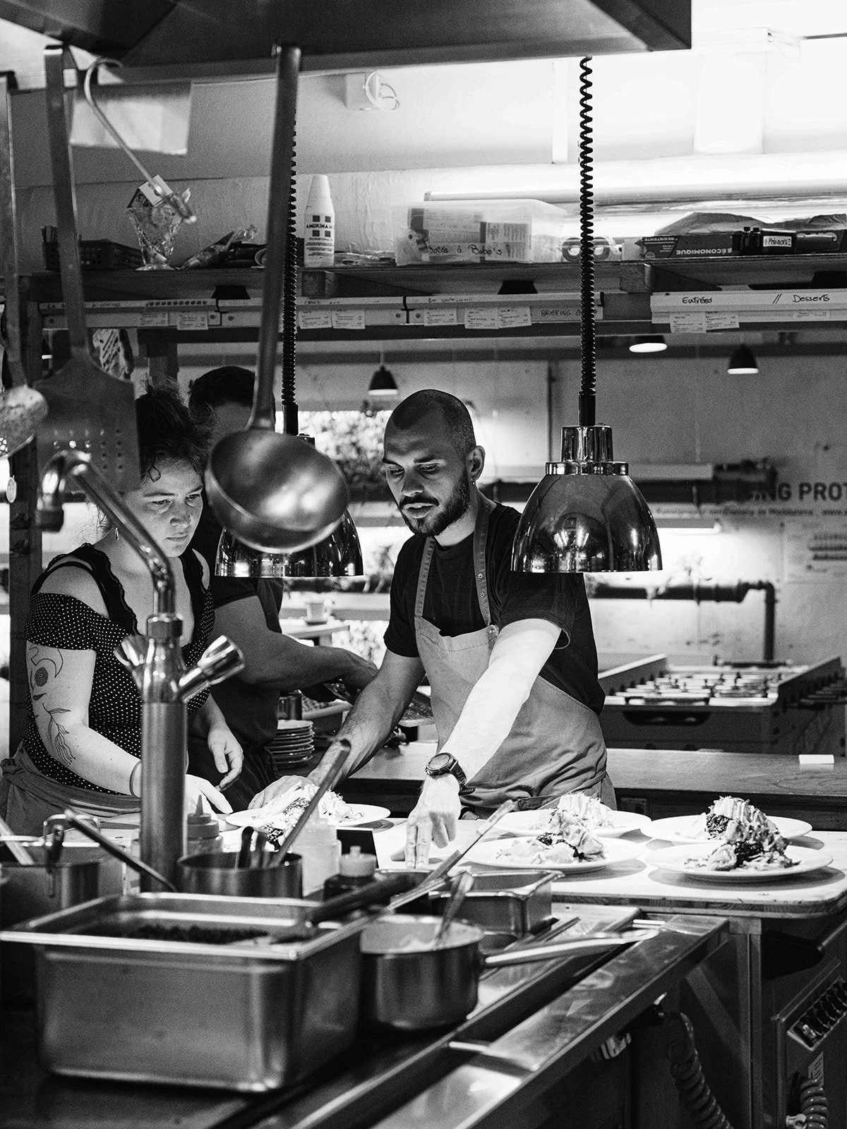 Deux cuisiniers travaillent ensemble dans une cuisine professionnelle, en train de préparer des plats. Un homme portant un tablier blanc et une montre, et une femme avec des tatouages, sont concentrés sur leur tâche.