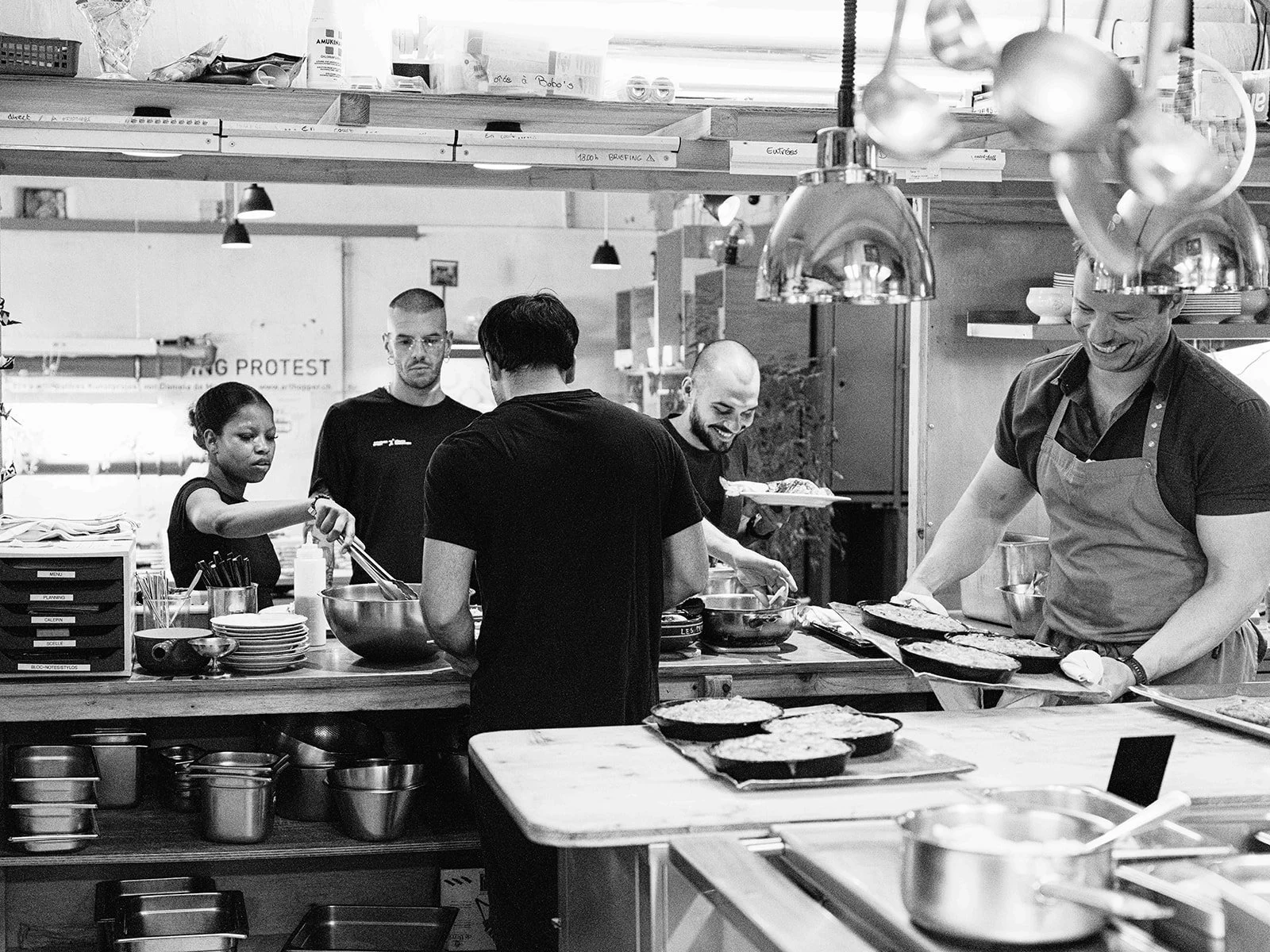 Groupe de personnes dans une cuisine de restaurant, certaines servant de la nourriture dans des plats, d'autres préparant ou organisant des plats, dans une ambiance de cuisine professionnelle