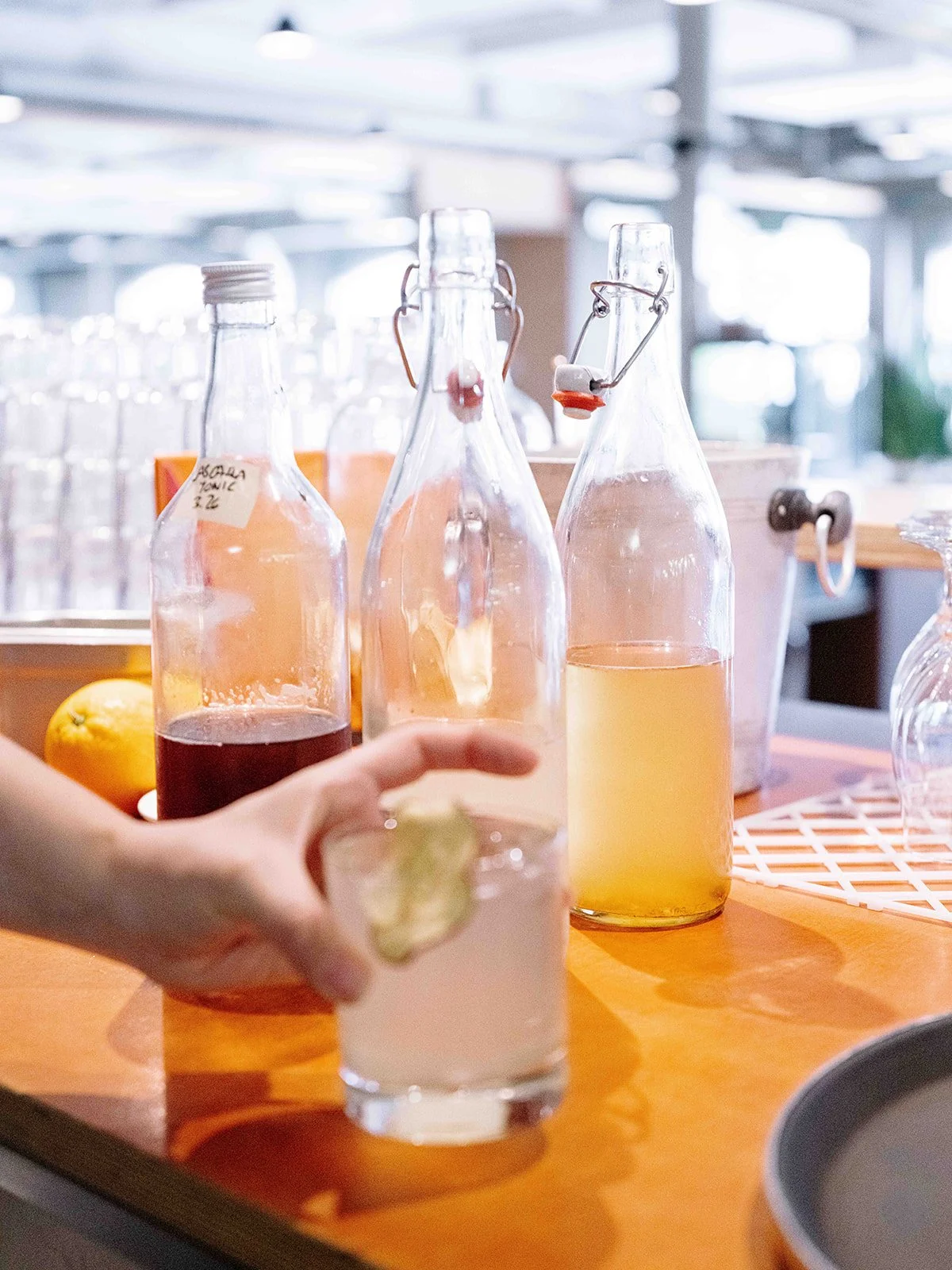 Bouteilles en verre contenant des liquides colorés sur une table en bois. Une main tenant un verre avec un citron vert. En arrière-plan, une cuisine avec des fenêtres lumineuses.