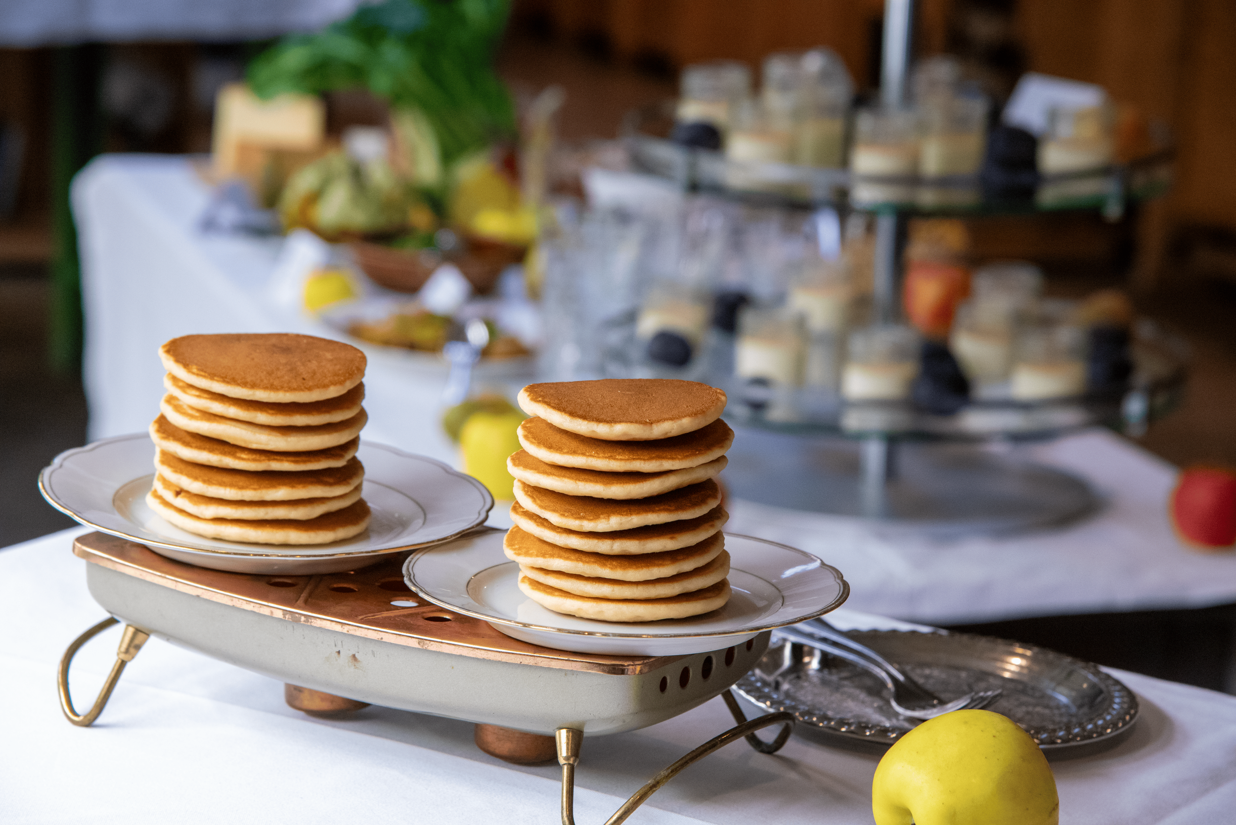 Une pile de pancakes dorés sur une assiette en porcelain, posé sur une table, avec un fond flou comprenant des aliments, une plante verte et un étagère avec des pots