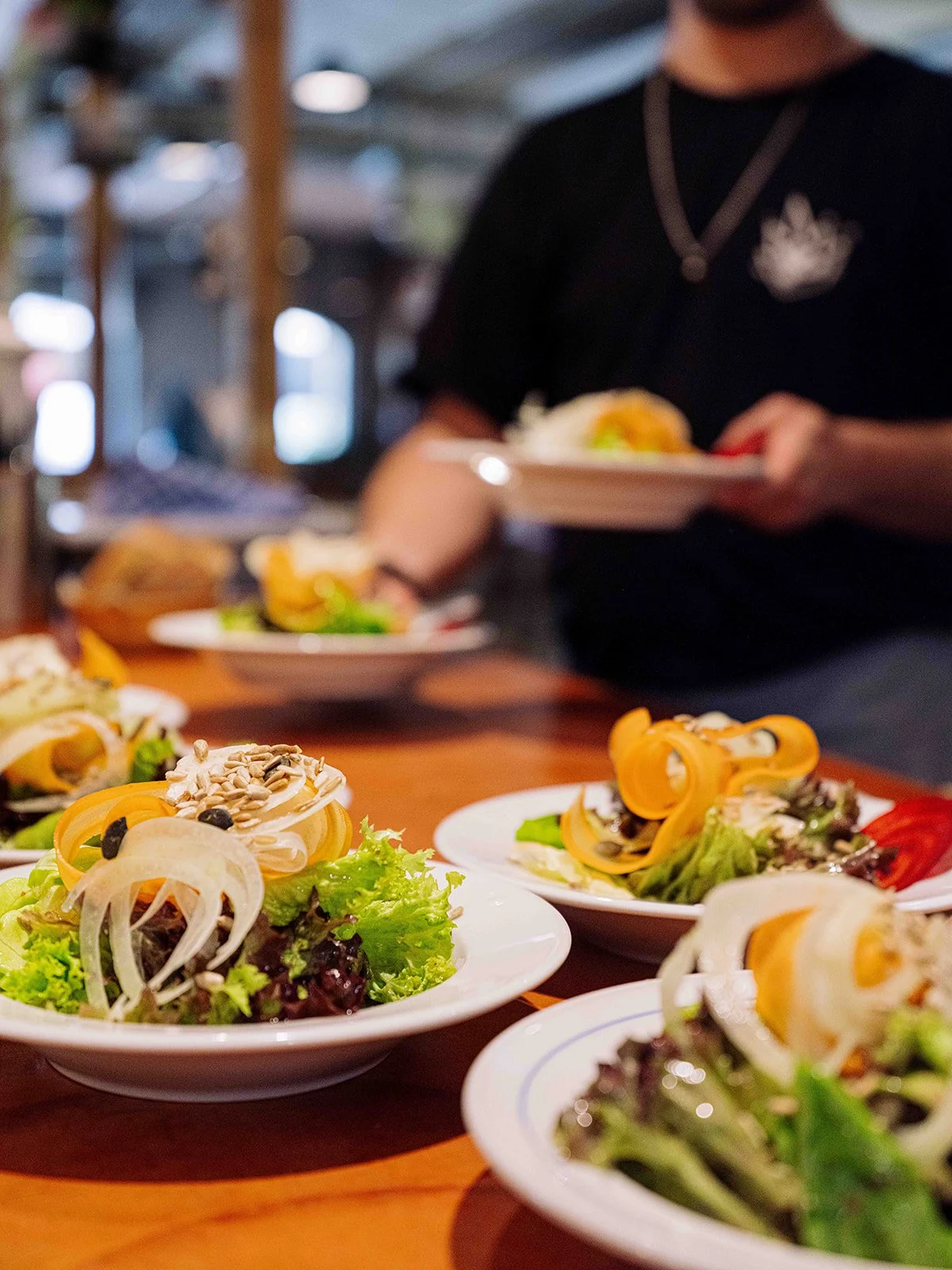 Salades servies sur des assiettes blanches garnies de légumes frais, graines et copeaux de fromage, posées sur une table en bois, dans un restaurant