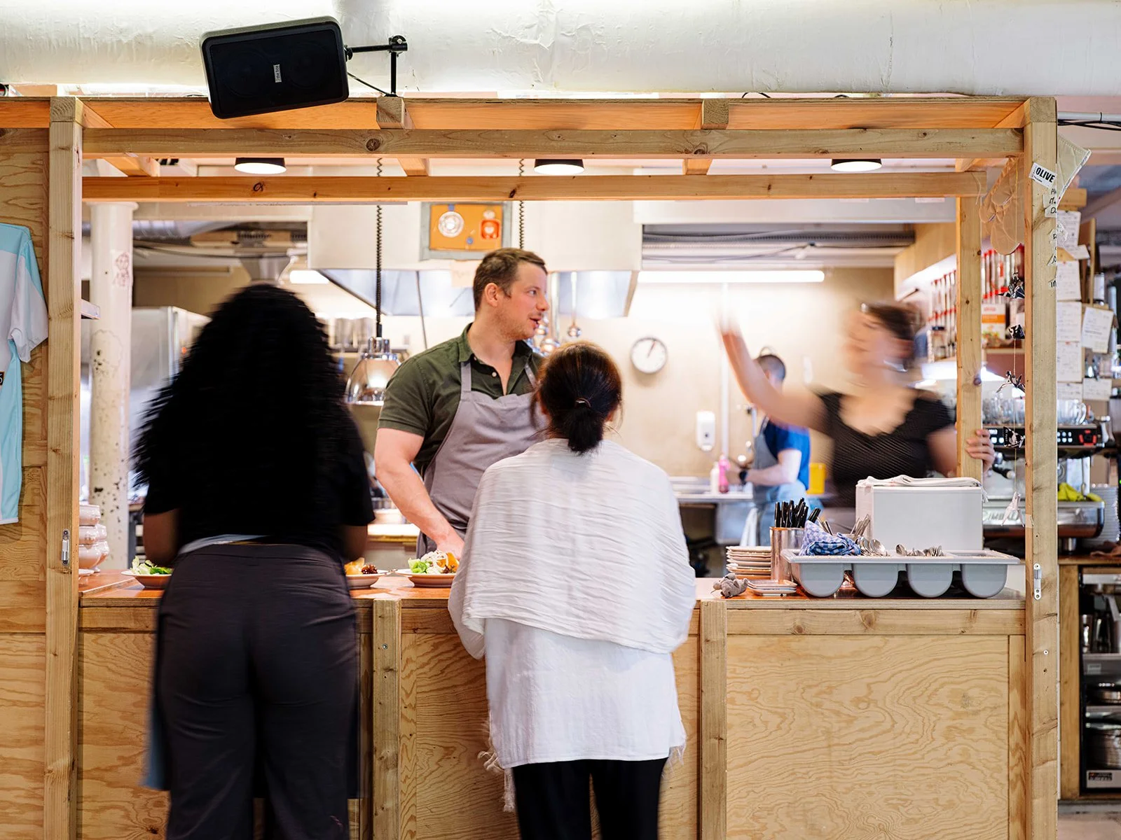 Une scène dans un restaurant où deux femmes sont assises au comptoir, discutant avec un homme derrière le bar, qui semble être le cuisinier ou serveur, tandis qu'une autre femme se tient à gauche, occupée avec ses plats.