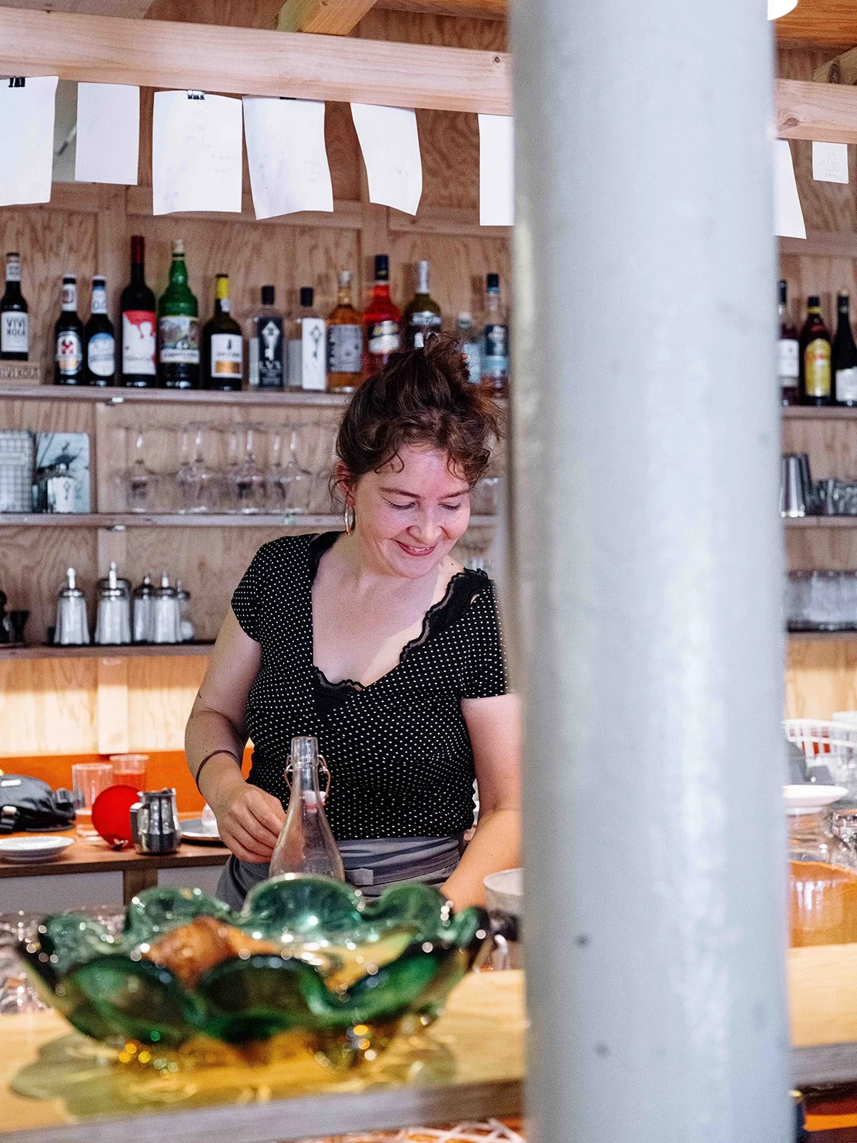 Une femme souriante dans une cuisine ou un bar, entourée de bouteilles d'alcool en arrière-plan, se préparant ou servant une boisson.