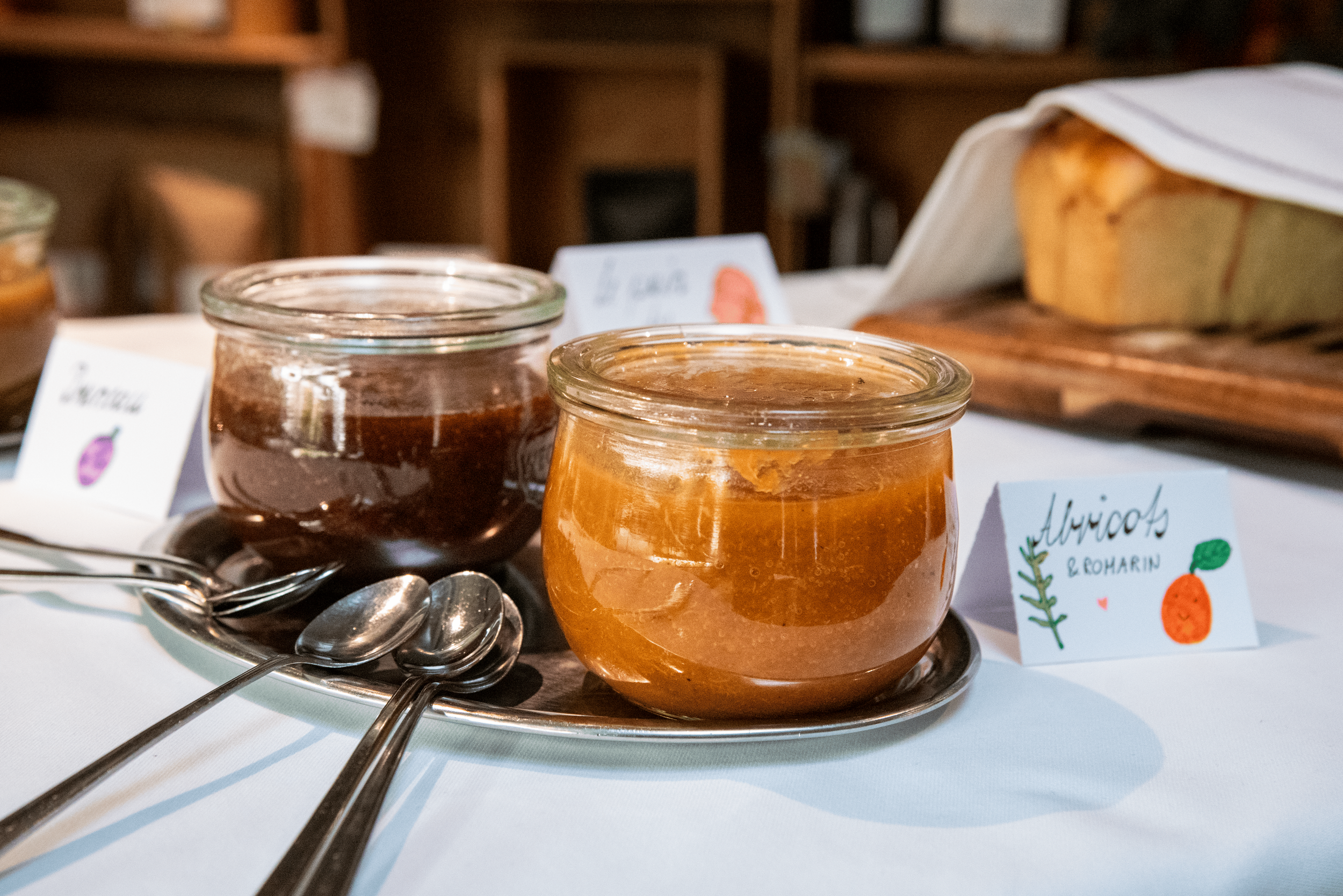 Petits pots en verre de confiture ou de gelée, placés sur un plateau en métal avec des cuillères en argent, devant des étiquettes colorées indiquant différents types de confitures ou gelées, dans un cadre intérieur