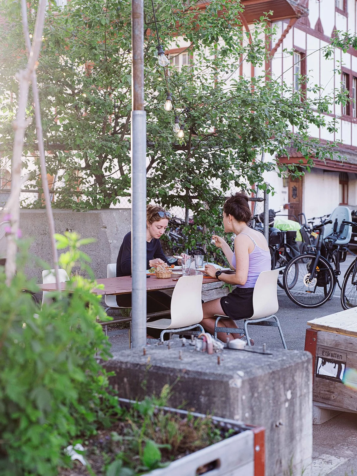 Deux femmes mangent à une table en extérieur sous un arbre, avec des vélos en arrière-plan, dans un cadre de quartier résidentiel.