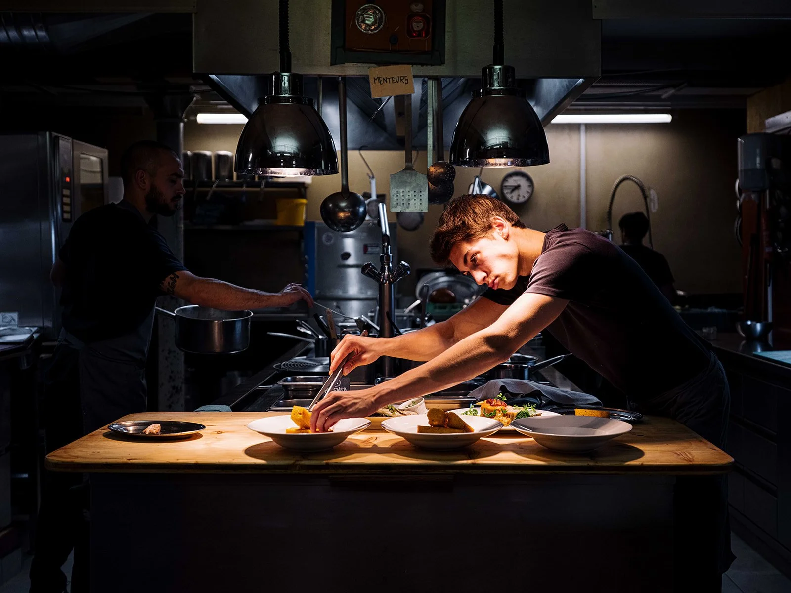 Deux jeunes hommes préparent des plats dans une cuisine professionnelle sombre, avec une lumière directe sur la surface de travail où ils disposent des assiettes de nourriture.