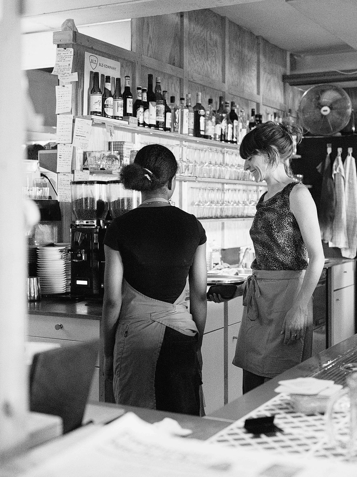 Deux femmes discutent derrière le comptoir d'un café ou restaurant, avec des bouteilles d'alcool et des verres en arrière-plan.
