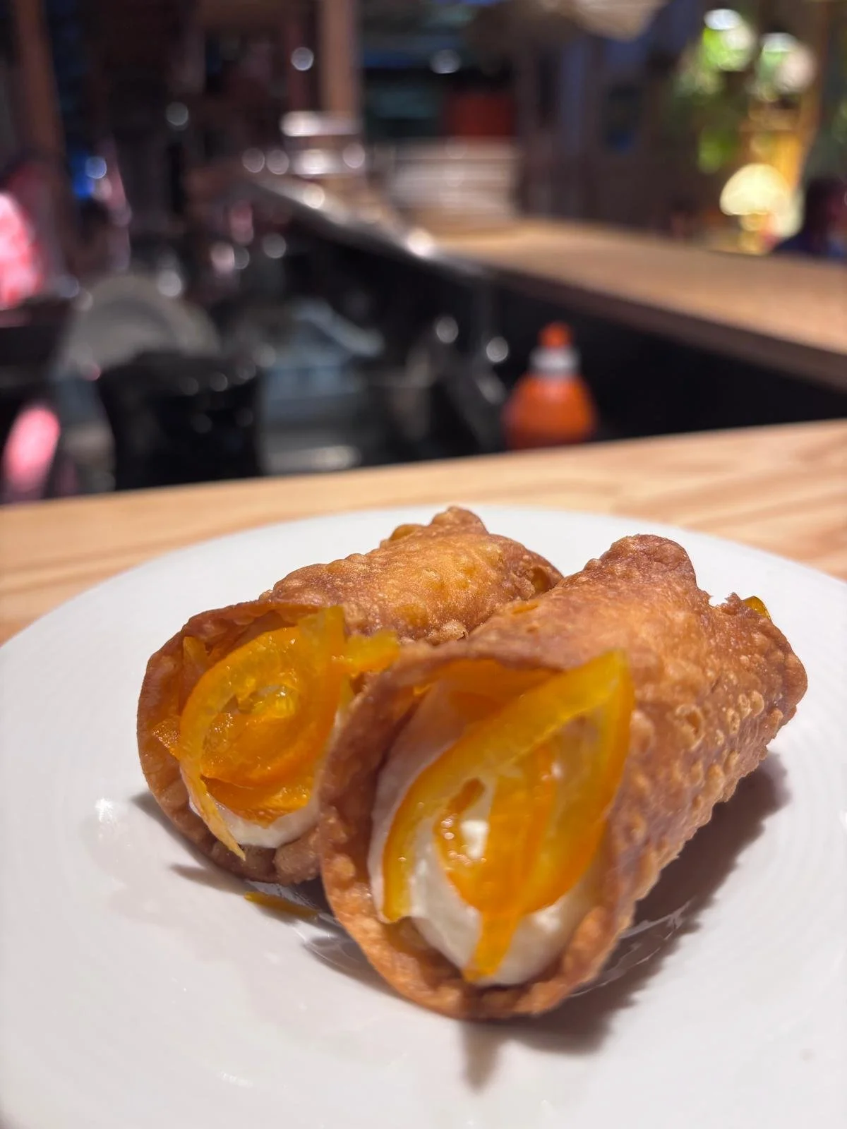 Deux rouleaux de printemps frits avec de la sauce piquante sur un plat blanc, en première plan, avec une cuisine floue en arrière-plan.