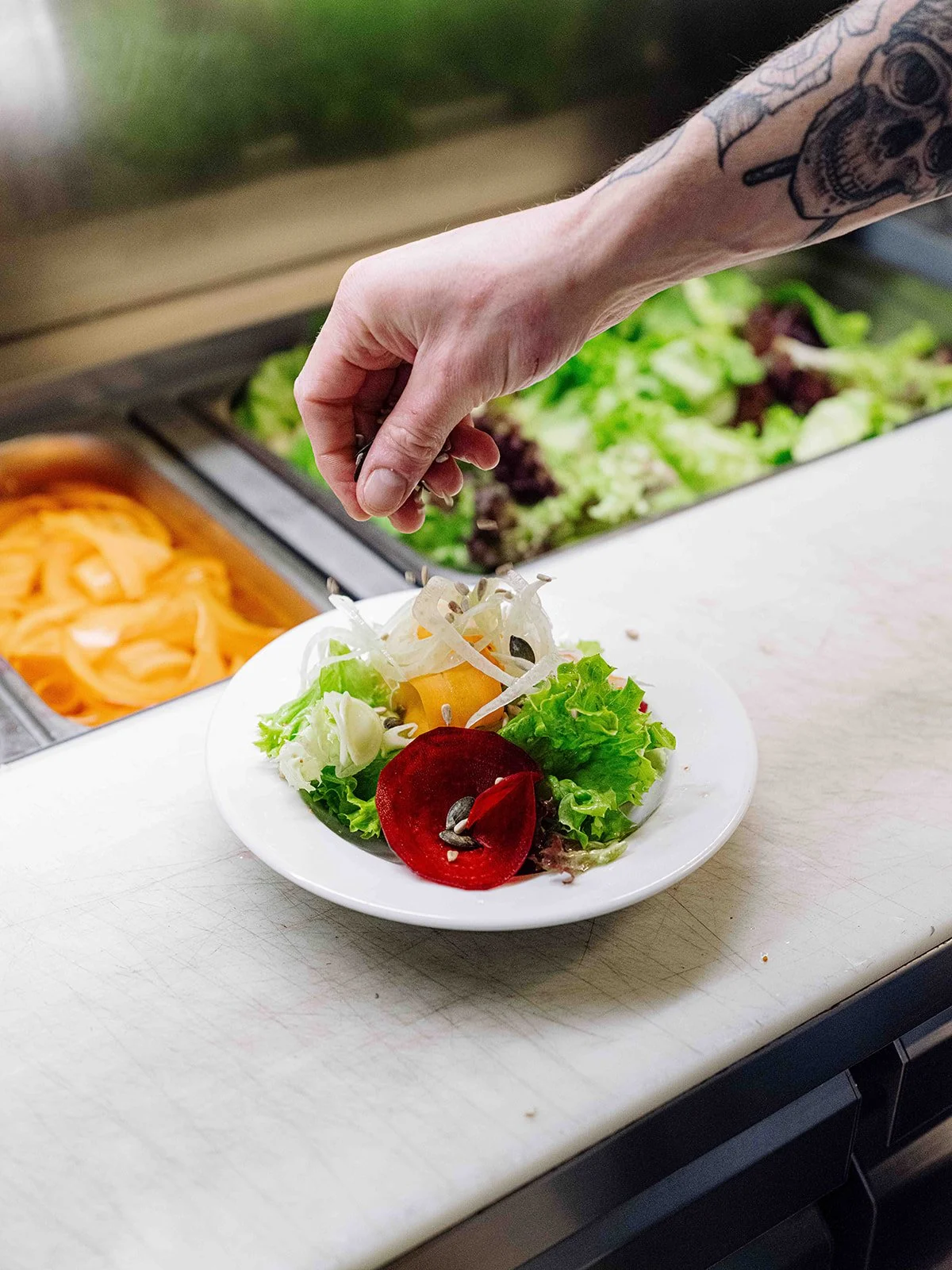 Une main contenant un tatouage verse des graines sur une salade composée sur une assiette blanche, avec une station de préparation de salades en arrière-plan.