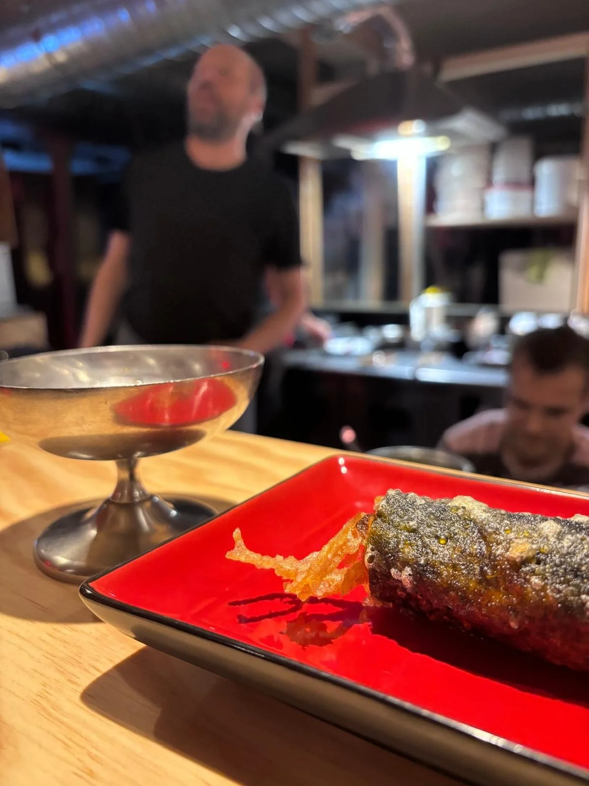 Une assiette rouge avec un rouleau de sushi et une friture, une coupe à dessert en métal vide, et deux personnes dans un restaurant japonais en arrière-plan.