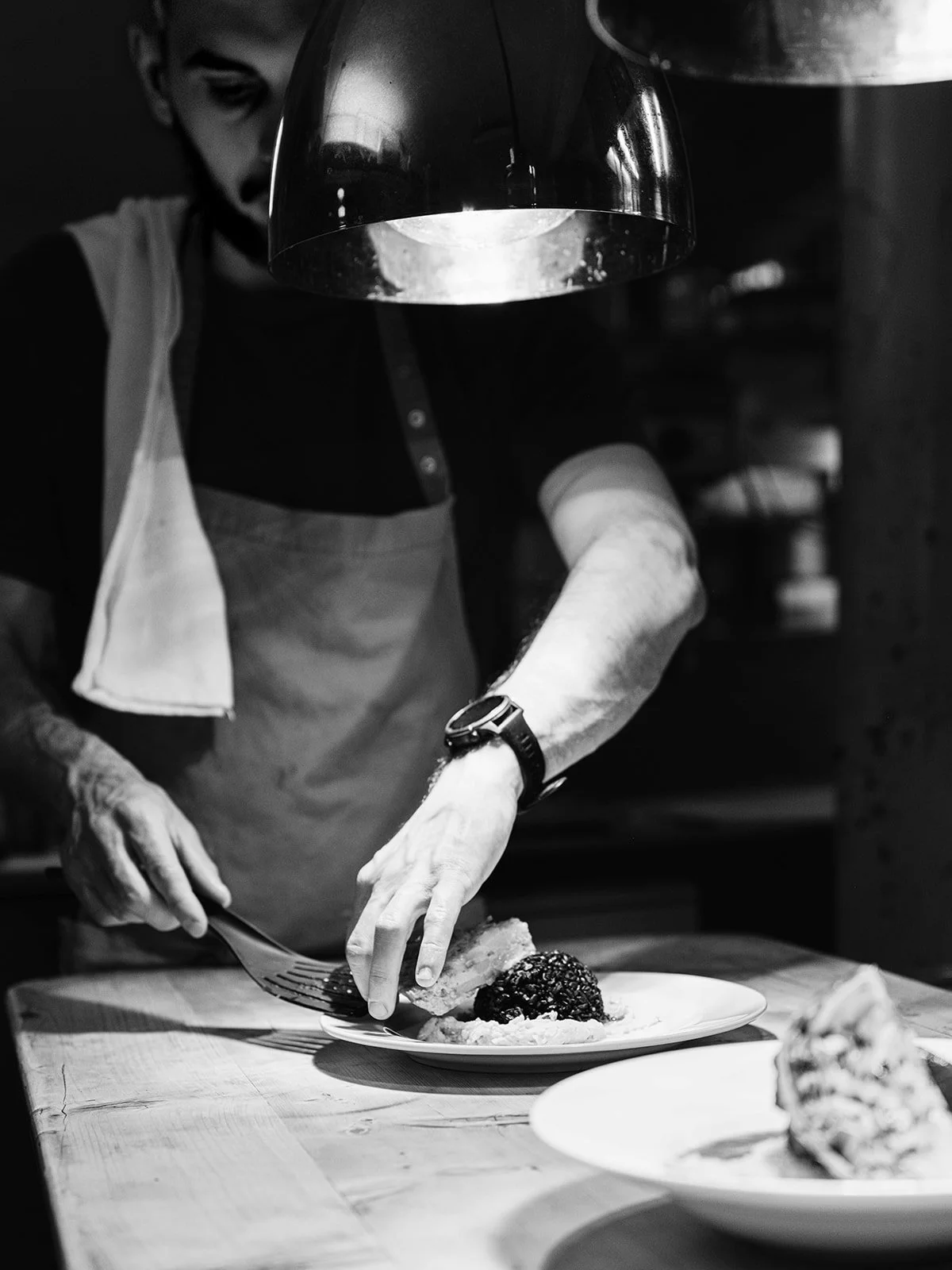 Un chef prépare un plat dans une cuisine en noir et blanc, portant un tablier et un smartwatch, en utilisant une fourchette.