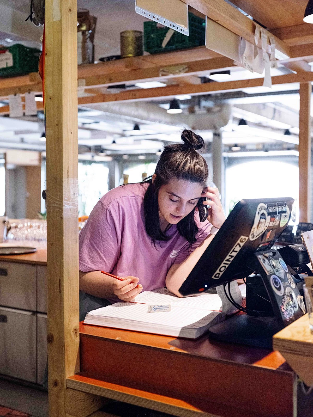 Jeune femme avec cheveux bruns en haut, vêtue d'un t-shirt rose, parlant au téléphone tout en écrivant dans un cahier dans un espace de travail à aire ouverte avec poutres en bois.
