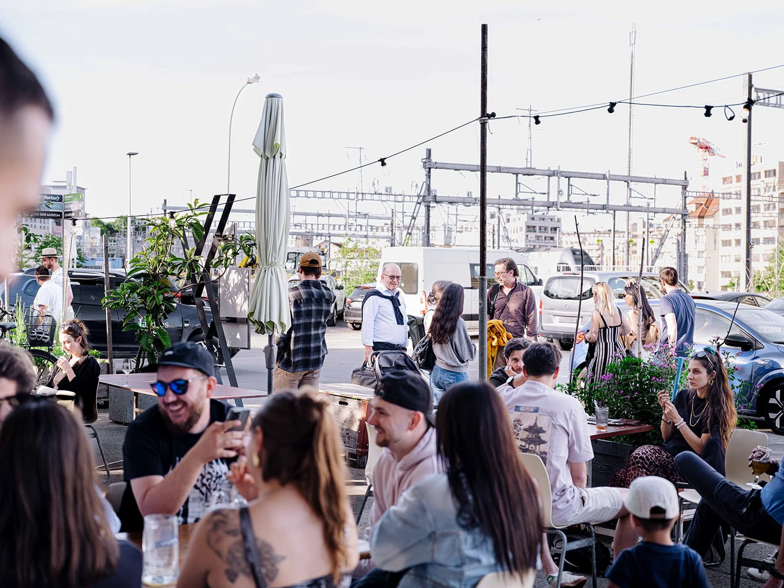 Une terrasse de restaurant ou café en extérieur avec plusieurs personnes assises ou debout, discutant et profitant du soleil, avec des voitures garées en arrière-plan et un ciel clair.