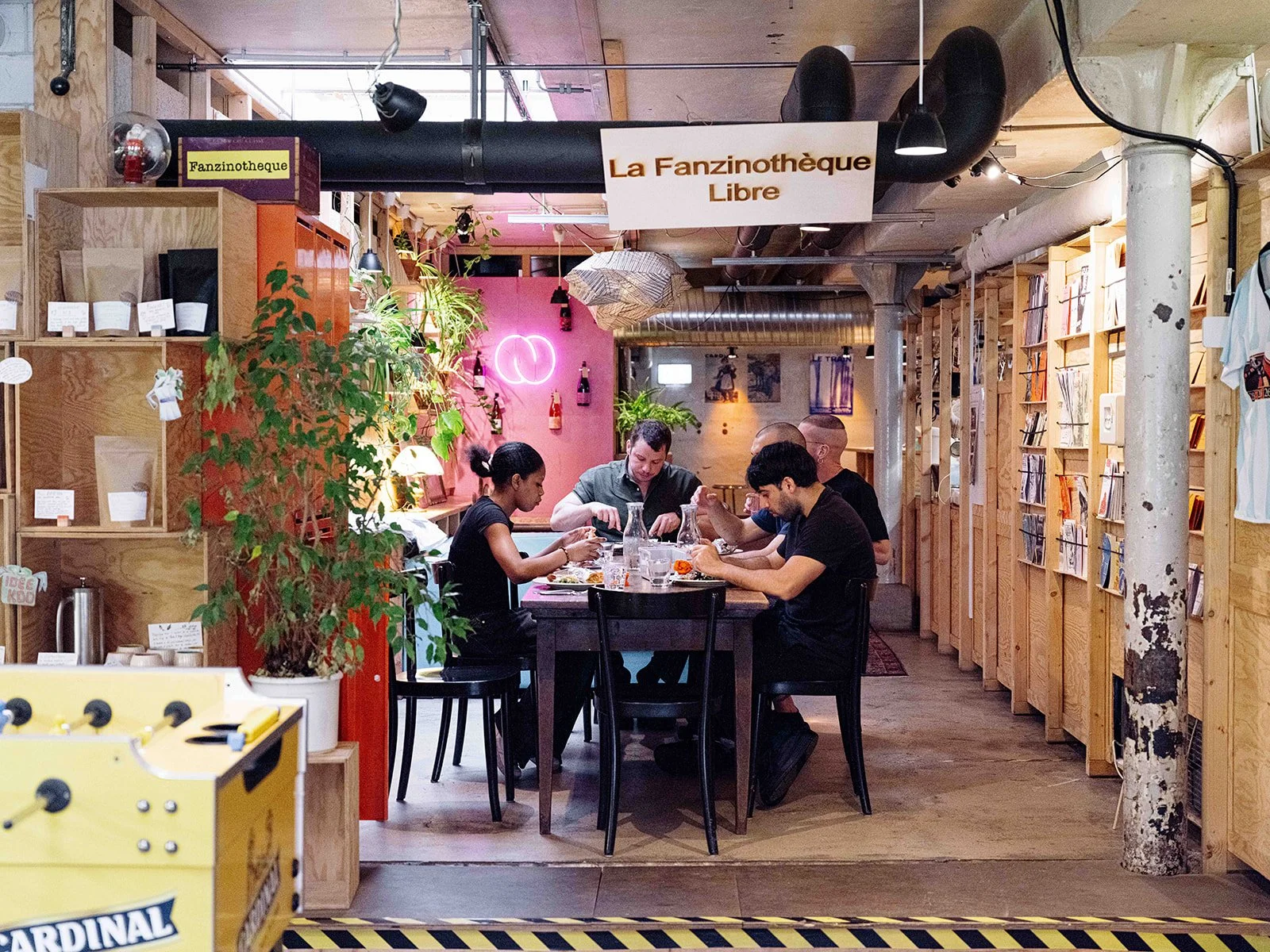 Groupe de cinq personnes assises autour d'une table dans un café ou un espace de coworking, en train de manger et de parler, décor intérieur avec étagères en bois, plantes et éclairage moderne.