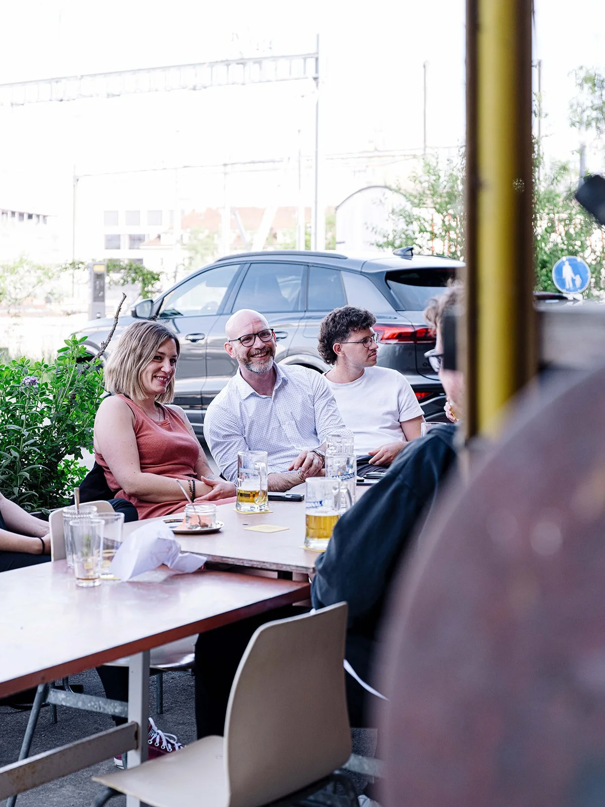 Un groupe de personnes assises autour d'une table à l'extérieur d'un restaurant, en train de boire des bières et de discuter, avec des voitures en arrière-plan.