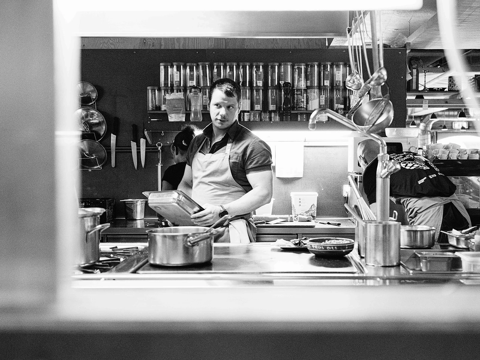 Cuisinier masculin dans une cuisine professionnelle, portant un tablier blanc et tenant une tablette, avec des casseroles et des ustensiles de cuisine autour.
