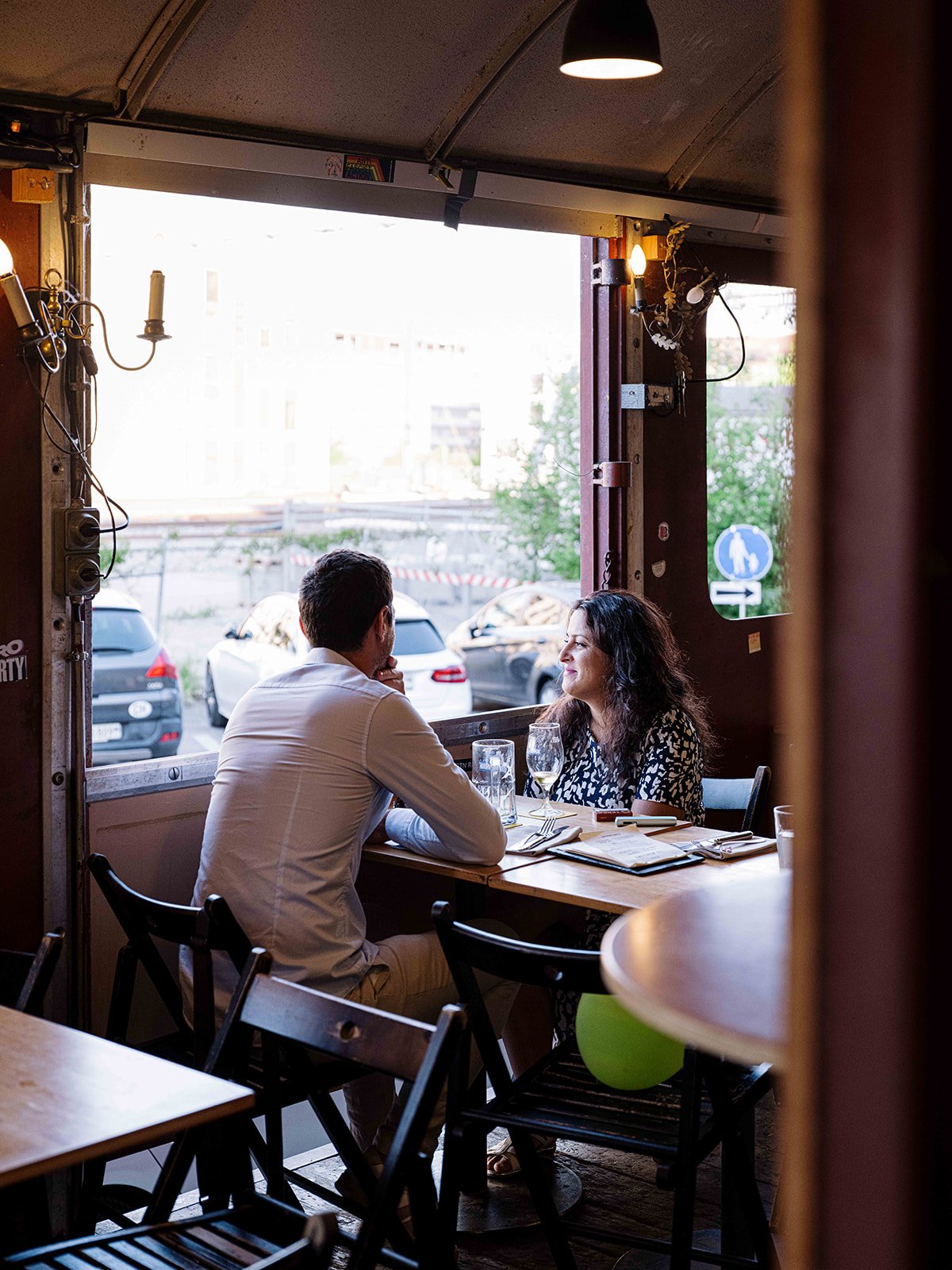 Deux personnes, un homme en chemise blanche et une femme avec des cheveux bouclés, discutent à une table dans un restaurant avec vue sur la rue parisienne.