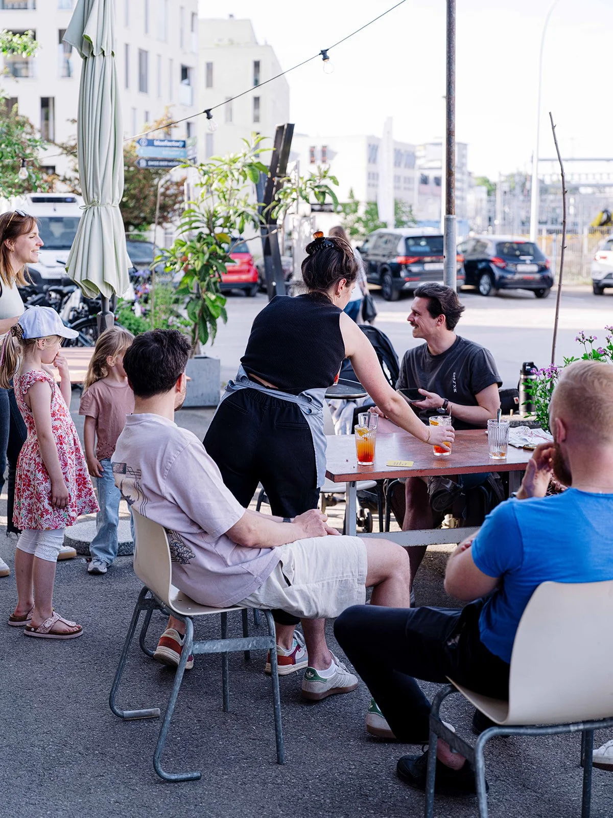 Groupe de personnes dans une terrasse de café en plein air, discutant et partageant des boissons, avec des enfants et des adultes, dans un environnement urbain avec des bâtiments en arrière-plan.