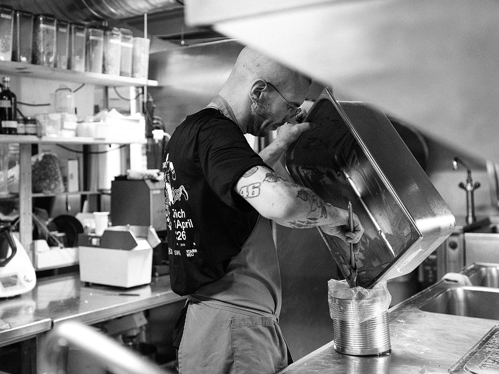 Un homme tatoué verse une substance dans une boîte en métal dans une cuisine ou un atelier. Il porte une chemise à manches courtes, un tablier, et des lunettes.
