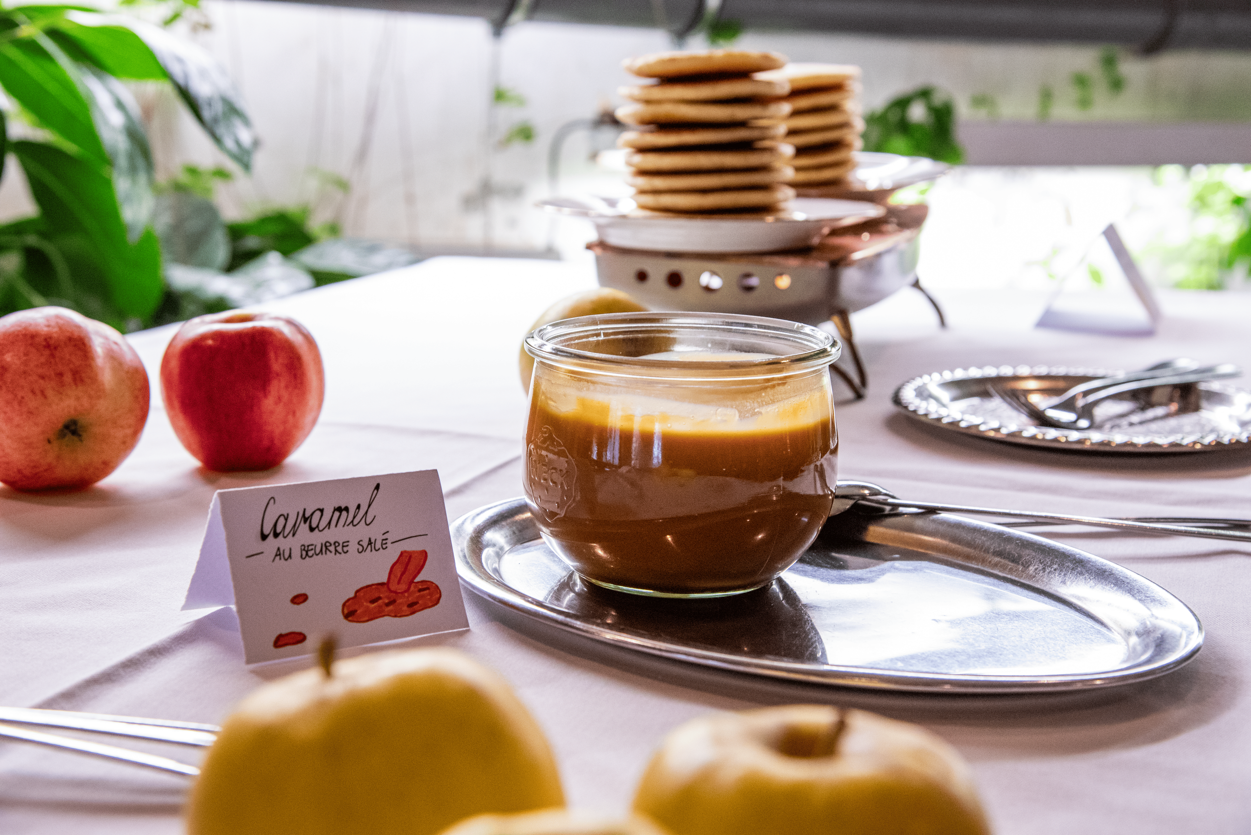 Un tableau avec des pommes rouges, un pot de caramel au beurre salé, une assiette avec des biscuits, et une petite plaque avec une illustration de biscuits et la mention "Caramel au beurre salé".
