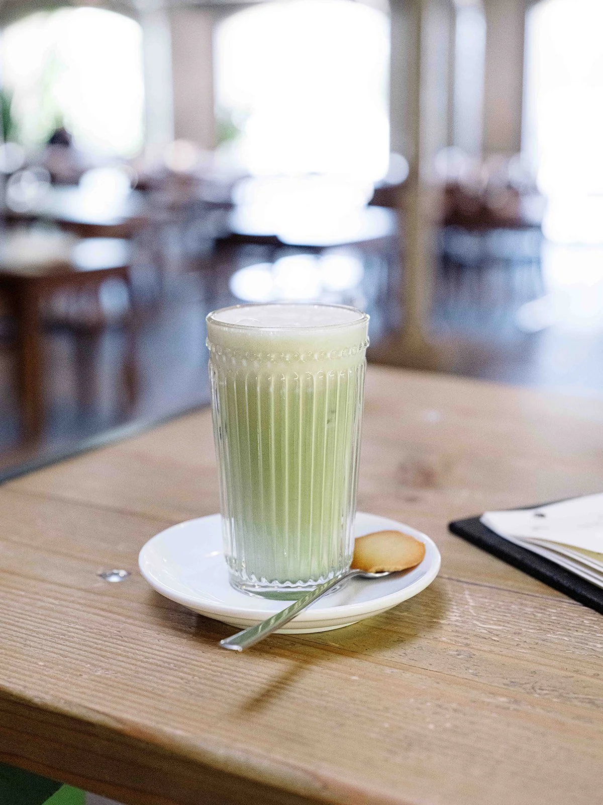 Un verre de boisson verte sur une assiette blanche avec une cuillère, posé sur une table en bois. En arrière-plan, une salle de café ou restaurant est visible mais floue.