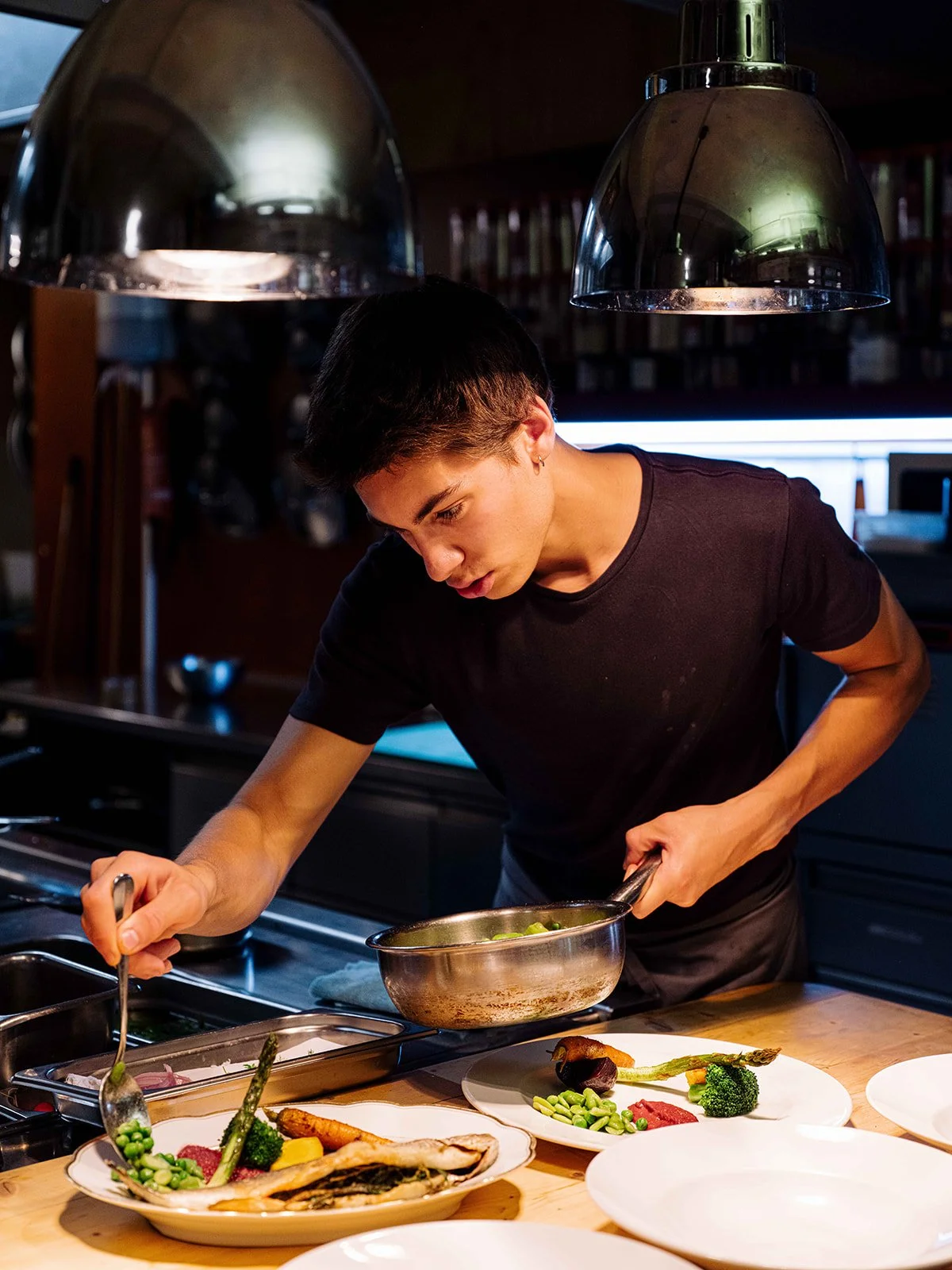 Jeune femme préparant un plat dans une cuisine moderne, entourée de plusieurs assiettes de légumes et poissons, sous des luminaires suspendus.