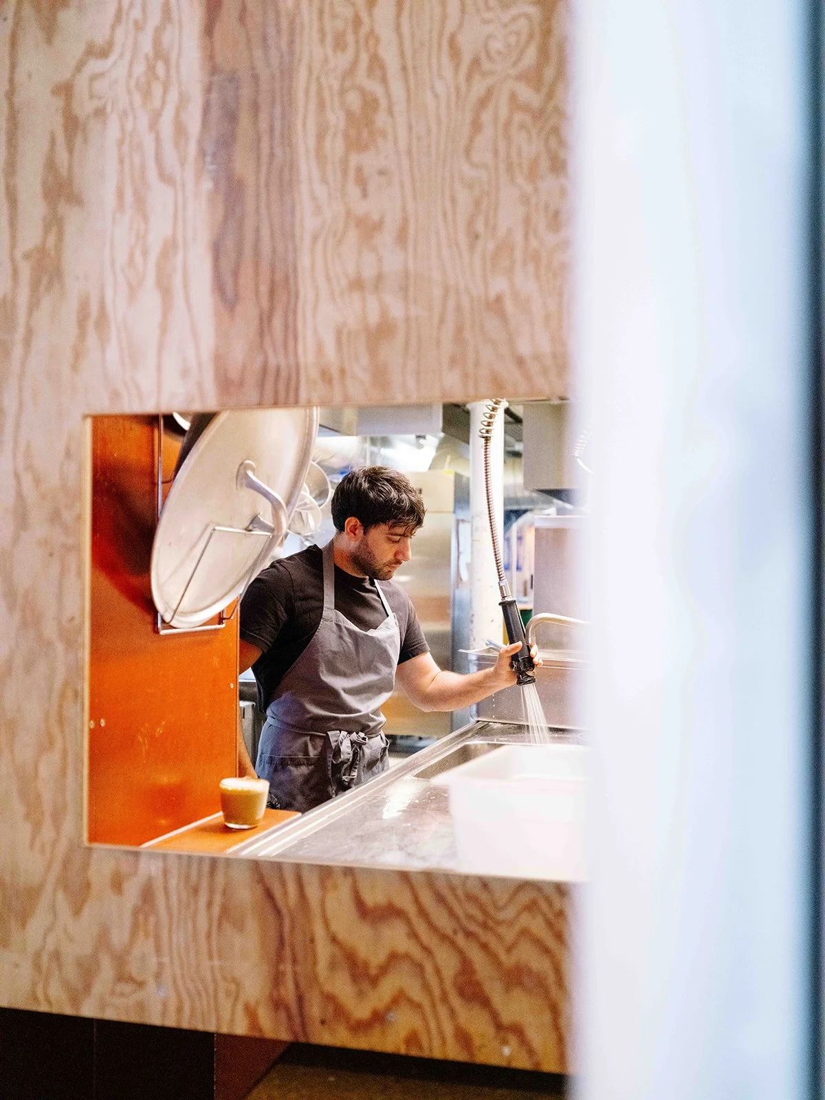 Un homme en tablier lave de la vaisselle dans une cuisine d'un restaurant vue à travers une ouverture dans un mur en bois.