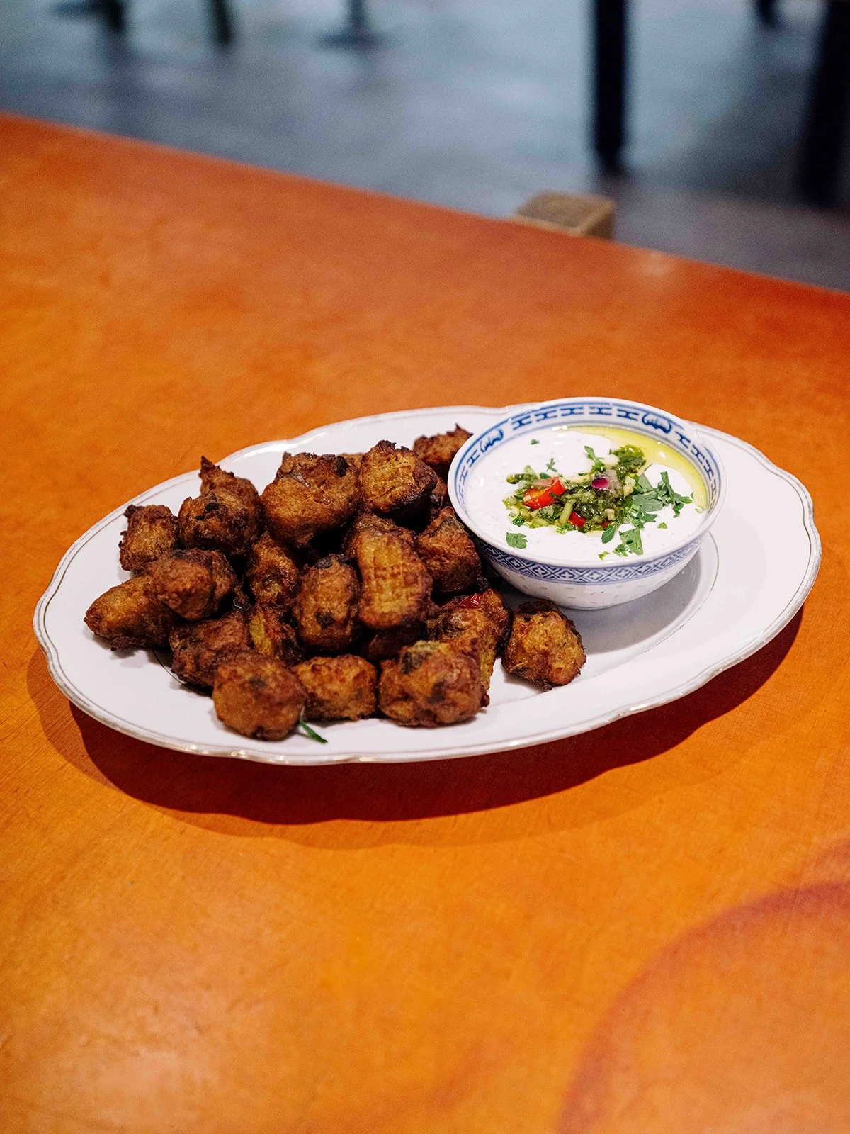 Gros plan sur un plateau de beignets frits avec une petite coupelle de sauce à côté, décorée de herbes et de petits morceaux de piment, sur une table en bois.