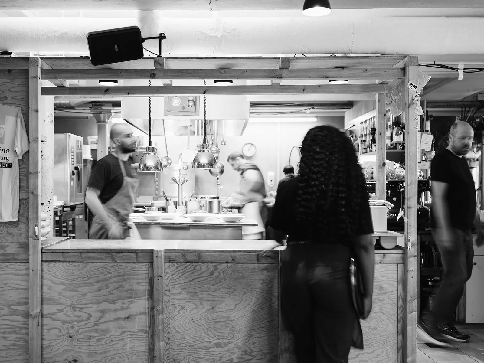 Une image en noir et blanc d'une cuisine dans un restaurant, avec plusieurs personnes, dont deux chefs en train de cuisiner et une personne qui attend devant le comptoir.