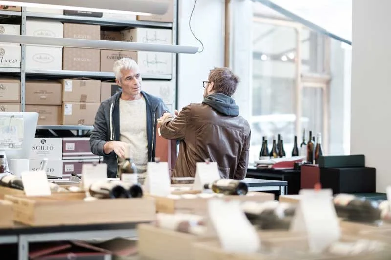 Deux hommes discutent dans un magasin de vin ou de spiritueux, avec des étagères et des bouteilles en arrière-plan.
