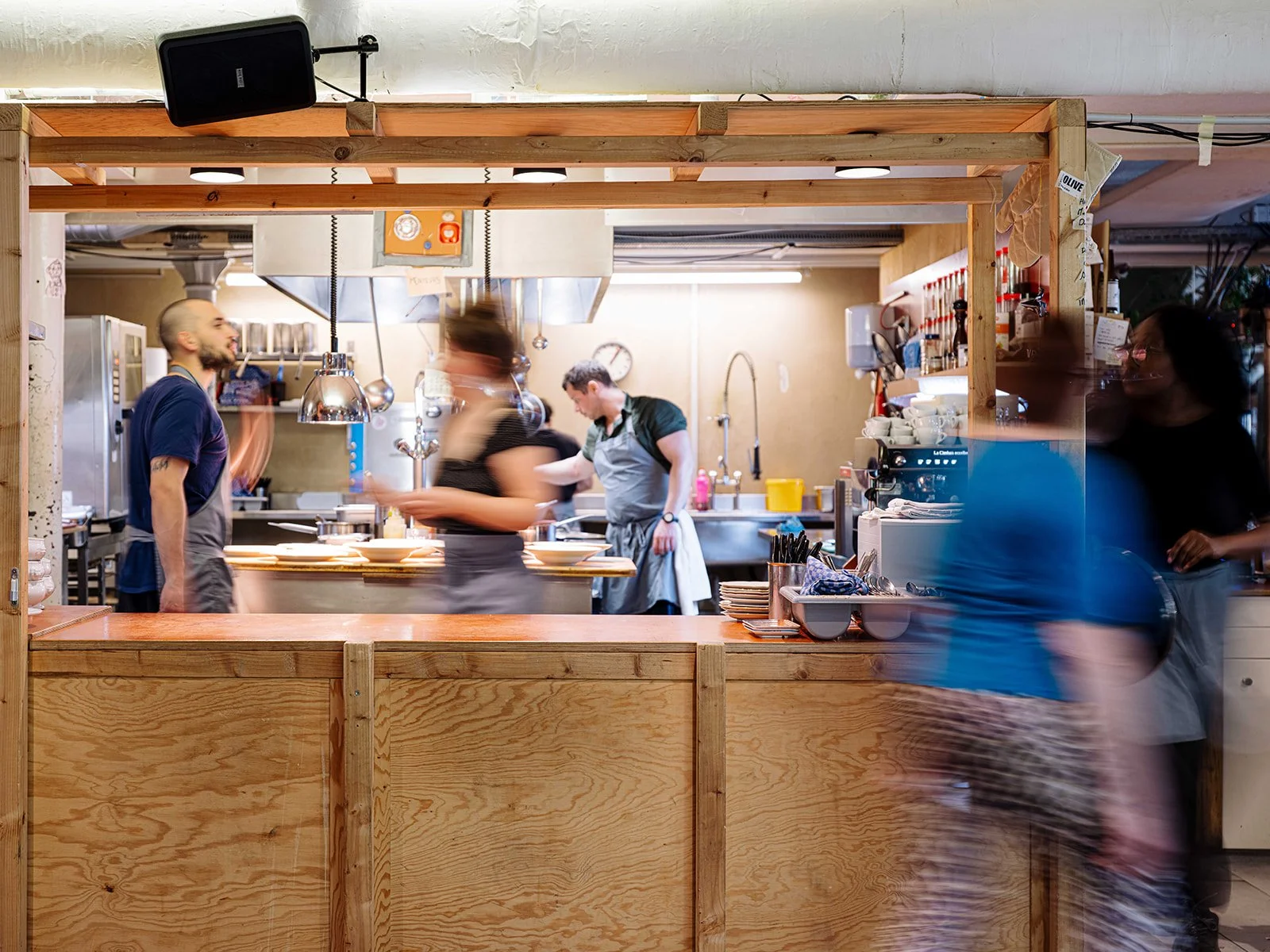 Vue de la cuisine d'un restaurant avec des chefs en action et des clients en arrière-plan, vue à travers un comptoir en bois.