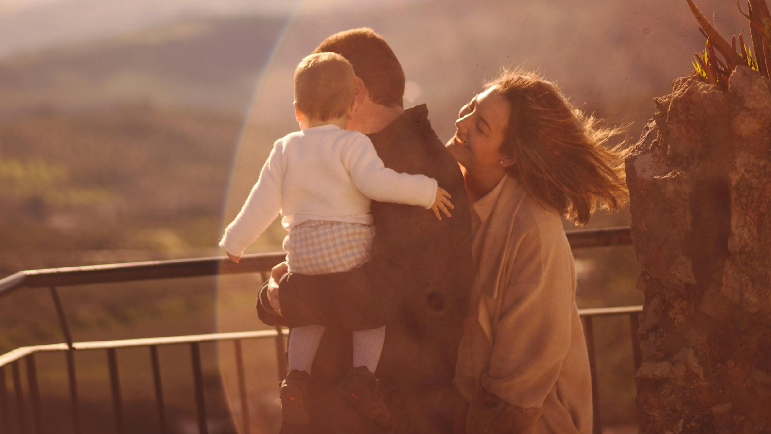 A woman holding a child in her arms while they share a joyful moment outdoors during sunset, with a rainbow and rocky landscape in the background.